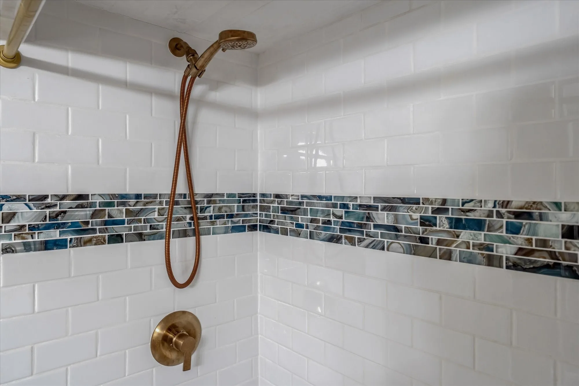 Shower enclosure featuring white subway tile surround, multi-tone glass mosaic accent band, and a bronze-finish shower head with handheld sprayer