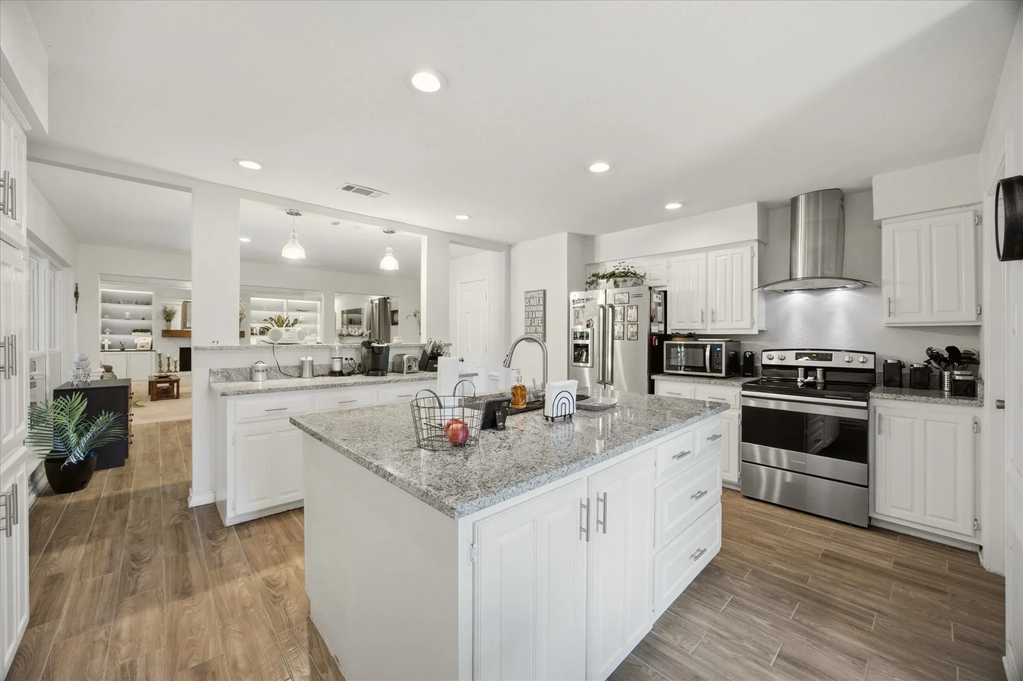 Open-concept kitchen featuring an expansive center island with a sink, white cabinetry, stainless steel appliances including a range and range hood, and wood-finish flooring