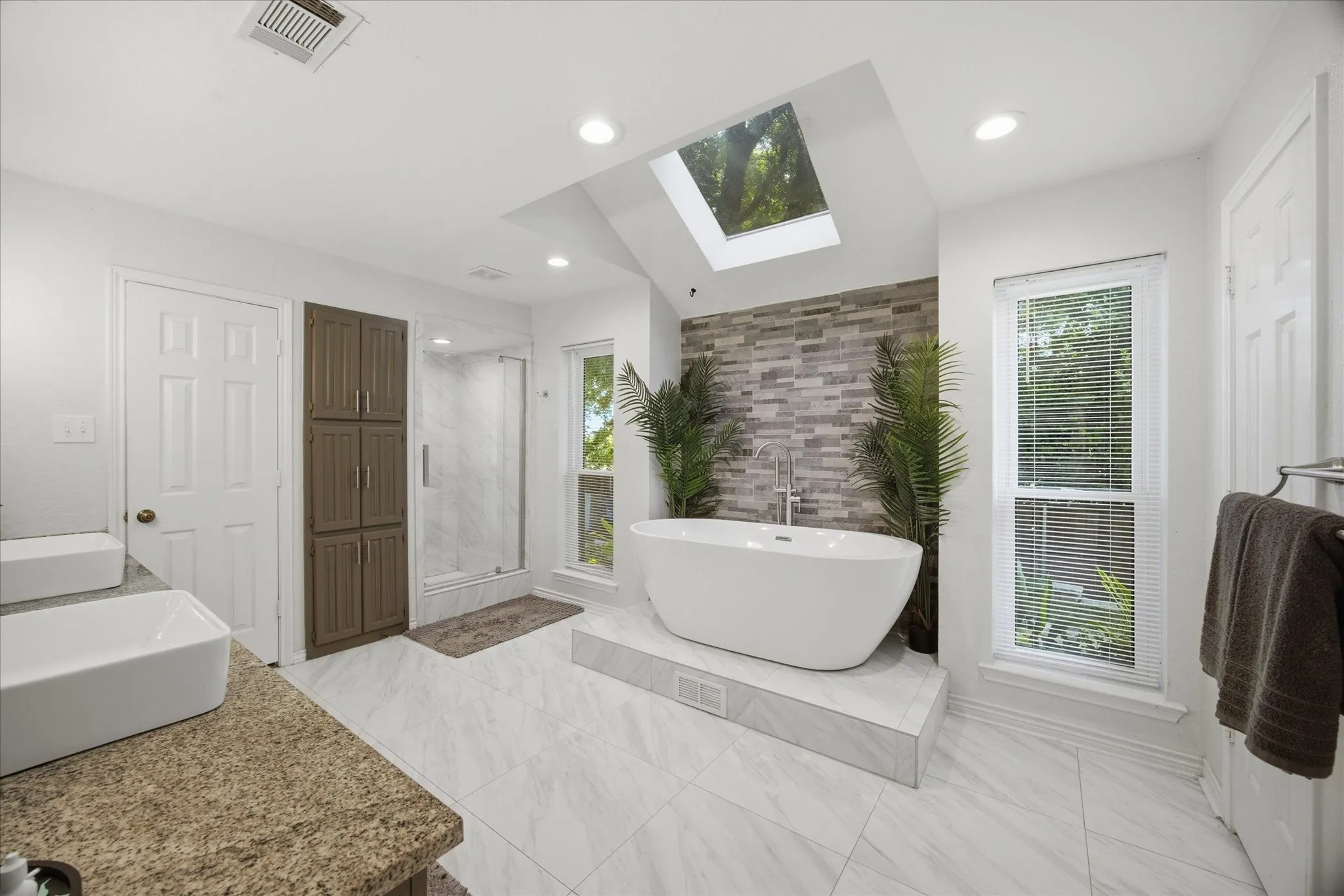 Spacious bathroom featuring a freestanding soaking tub on a raised platform, a stacked stone accent wall, a glass-enclosed shower, a double vanity with vessel sinks, and a skylight