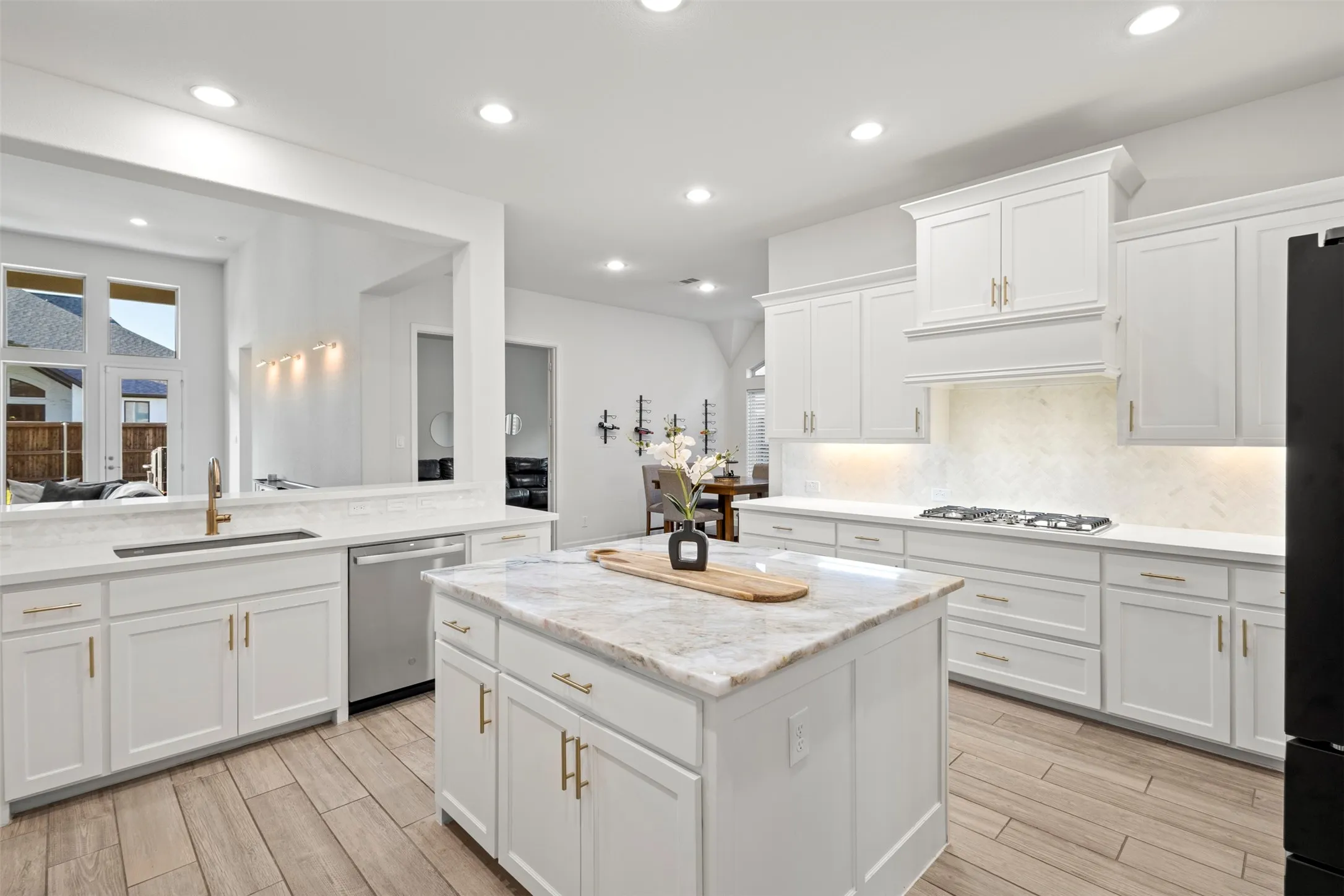 Contemporary kitchen featuring a large central island with a stone countertop, white cabinetry with gold-tone hardware, wood-finish flooring, a stainless steel dishwasher, and a gas range with an integrated hood