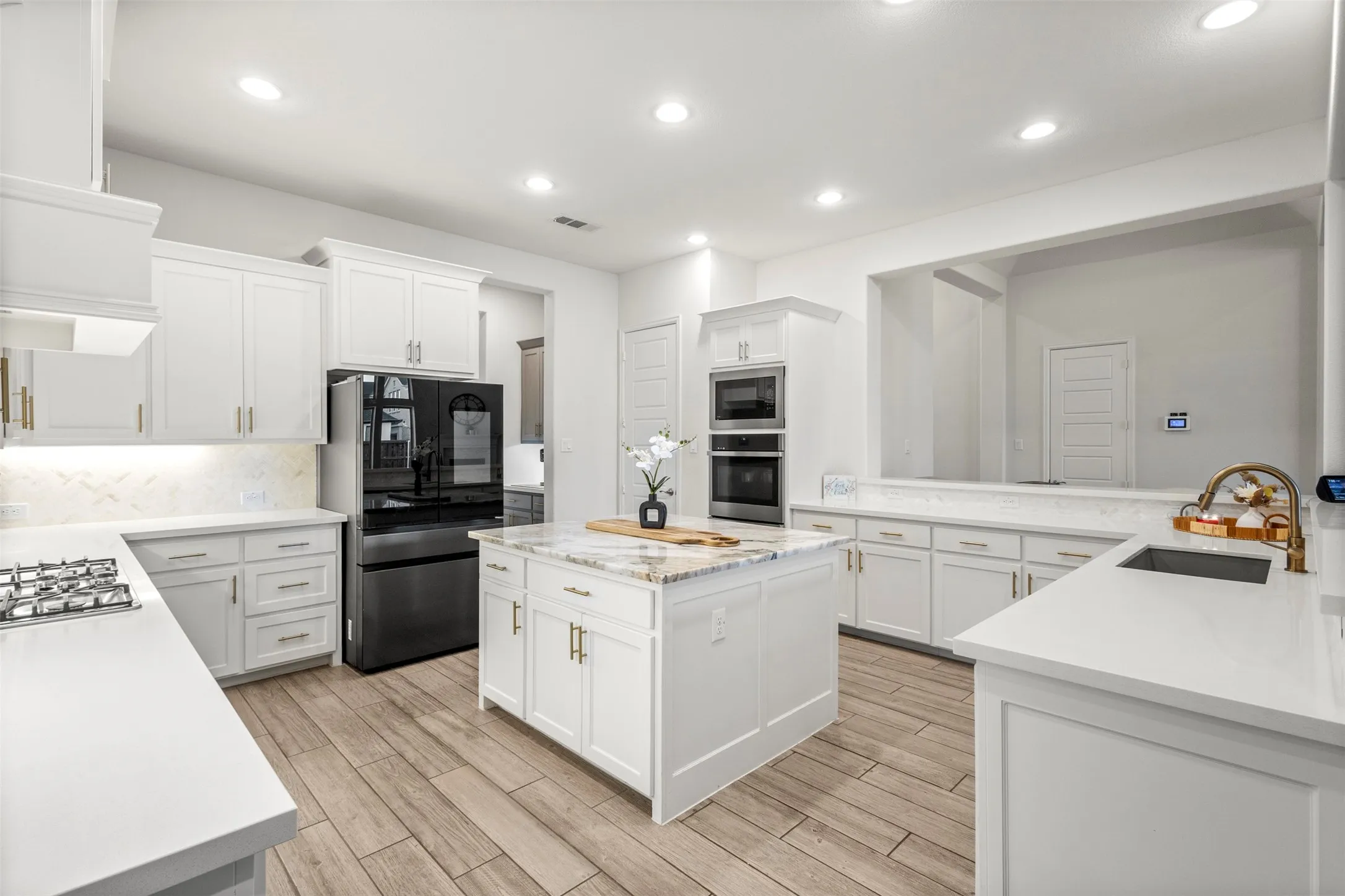 Gourmet kitchen featuring a central island with a granite countertop, white cabinetry with brass hardware, wood-finish flooring, built-in double ovens, and a gas cooktop