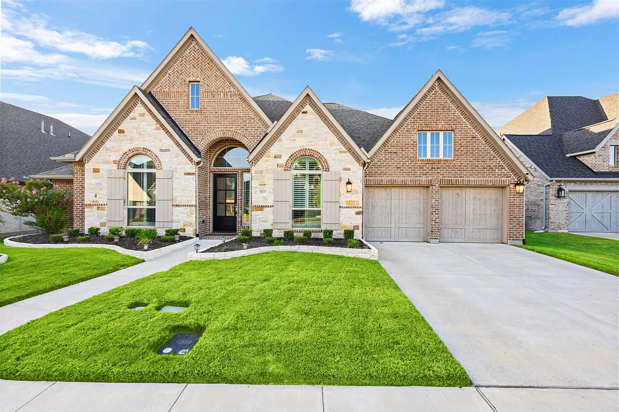 Brick and stone facade featuring arched entryways, a two-car garage with wood-finish doors, prominent gables, and a manicured lawn with a concrete walkway