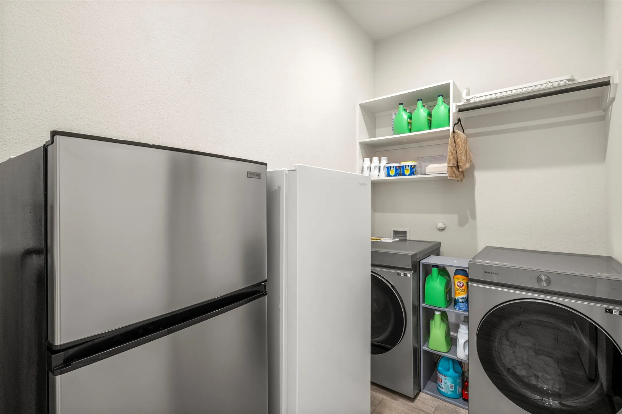 Dedicated utility space featuring a stainless steel refrigerator, washer and dryer appliances, white wall shelving, and an integrated hanging rod
