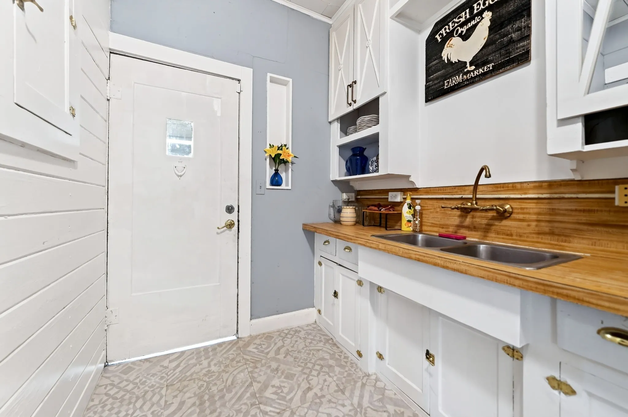 Double basin sink with a wall-mounted brass faucet, wood-finish countertops, white cabinetry with brass hardware, and patterned flooring