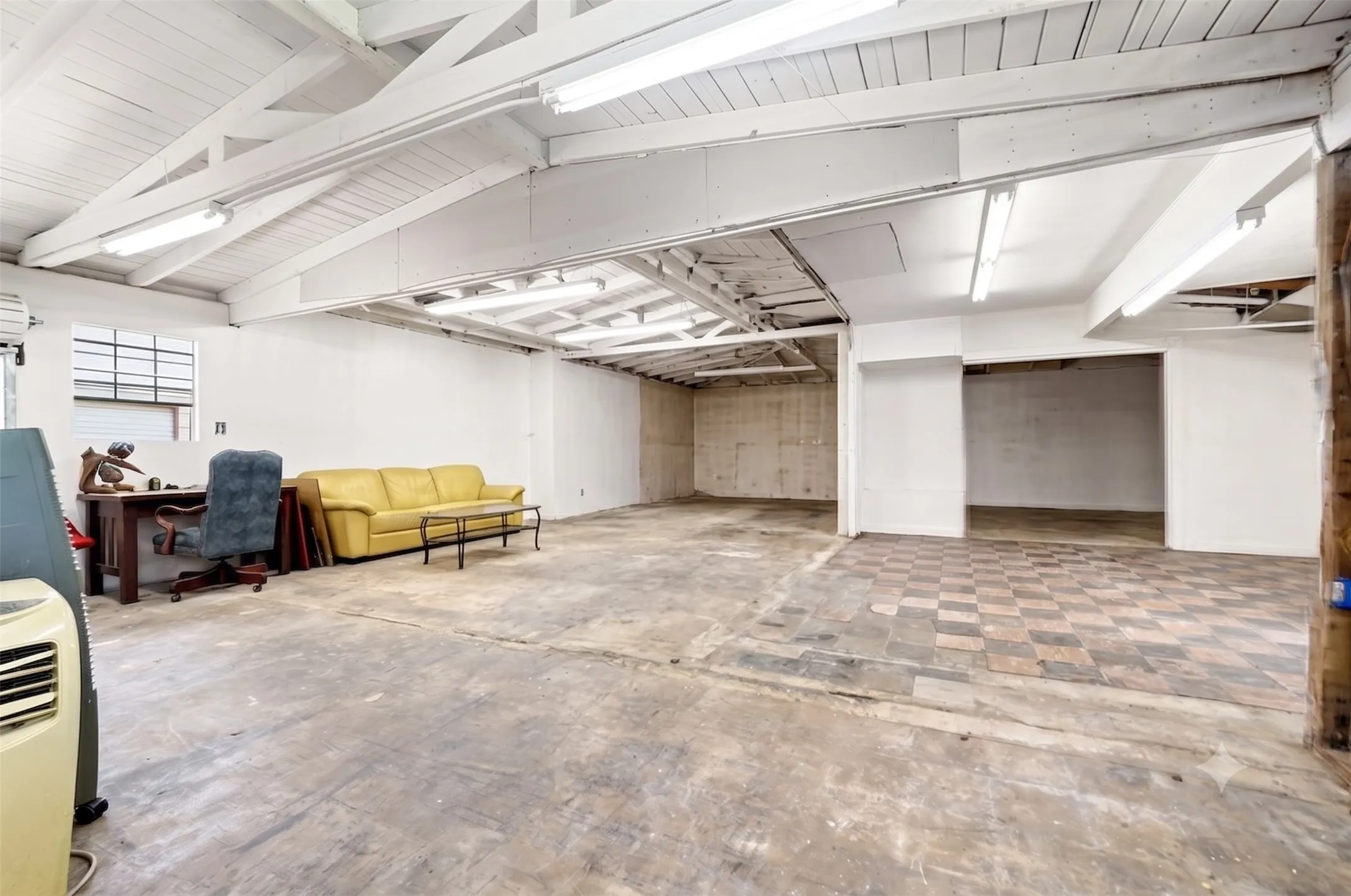 Spacious interior featuring exposed white ceiling beams, fluorescent lighting, a large window, and a combination of concrete and checkerboard tile flooring