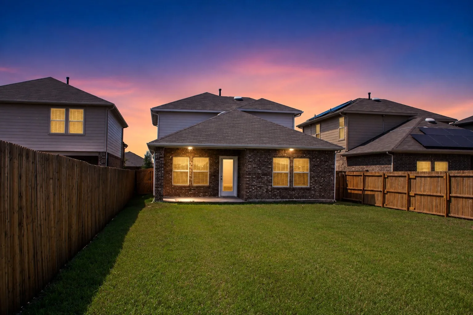 Expansive backyard featuring a manicured lawn, full privacy fencing, a brick exterior with multiple windows, and a paved patio area