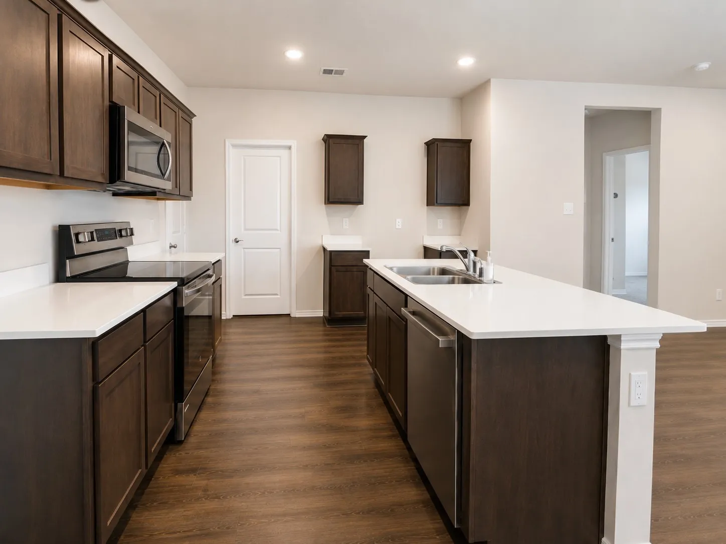 Spacious kitchen featuring dark wood cabinetry, white countertops, stainless steel appliances including a range and microwave, a kitchen island with an undermount double sink, and wood-finish flooring