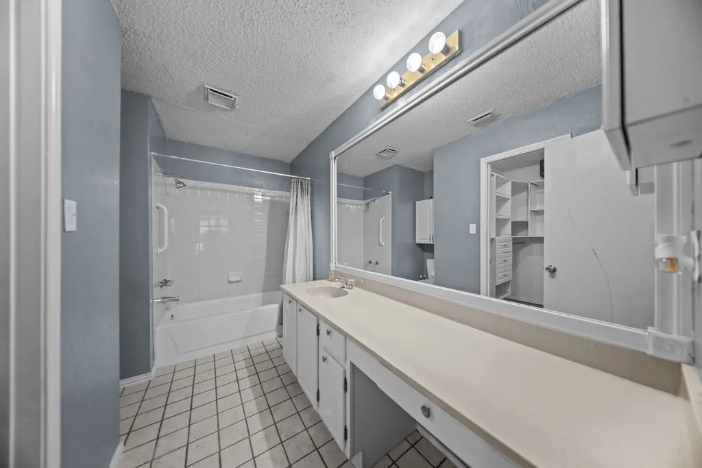 Bathroom featuring an extended vanity with dual sinks, a large wall-mounted mirror, a tub/shower combination with grab bars, and tiled flooring