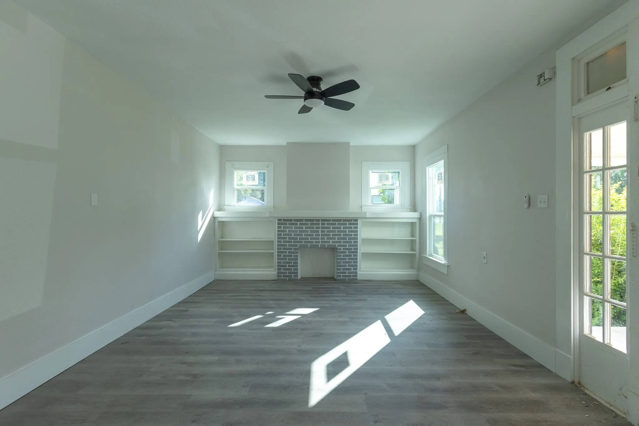 Spacious interior room featuring wood-finish flooring, a ceiling fan, and a fireplace with a light-colored brick surround