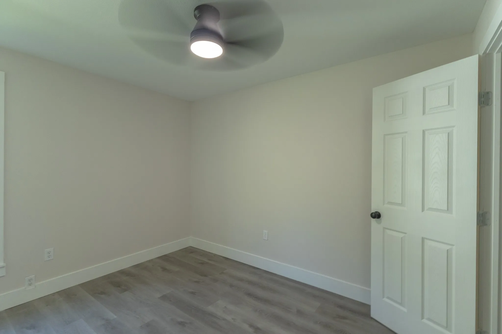 Bedroom featuring light gray walls, wood-finish flooring, a modern ceiling fan with integrated lighting, white baseboards, and a paneled interior door with a black knob
