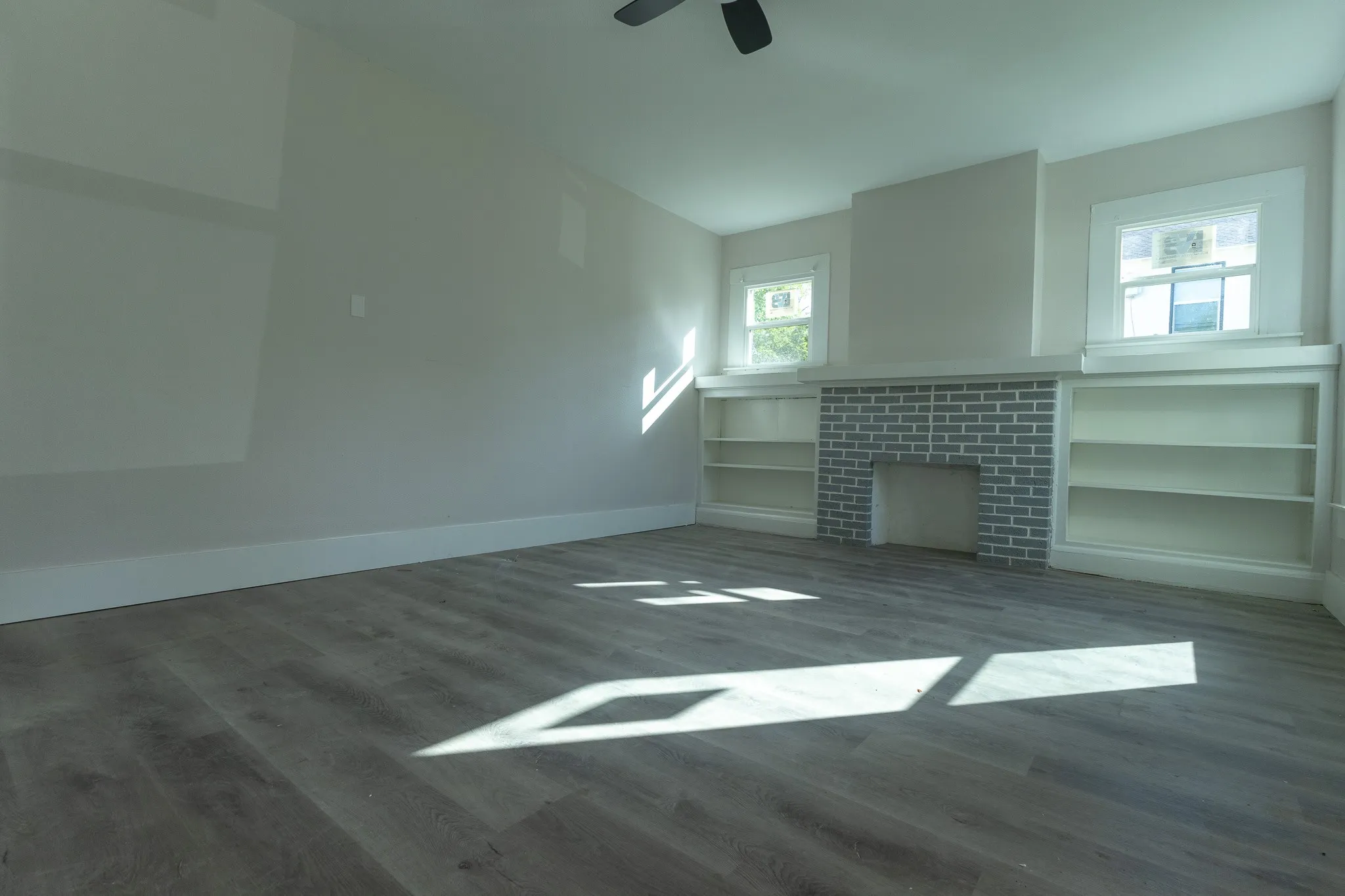 Spacious room featuring wood-finish flooring, a brick fireplace, twin windows, and built-in shelving