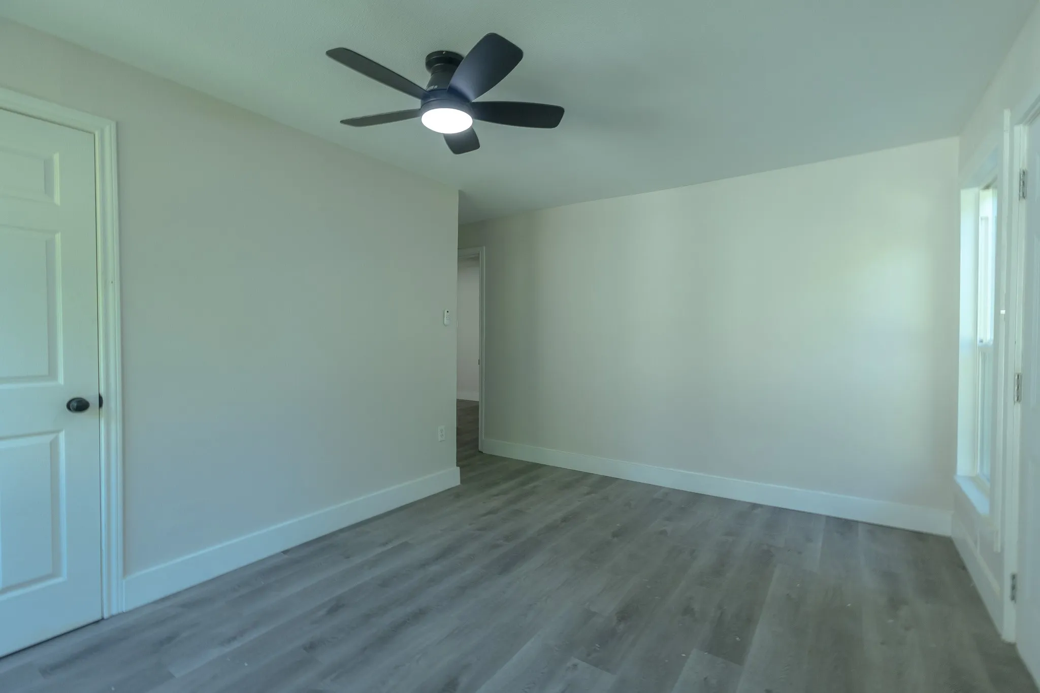 Spacious room featuring wood-finish flooring, a contemporary ceiling fan with integrated lighting, clean white baseboards, and a paneled interior door
