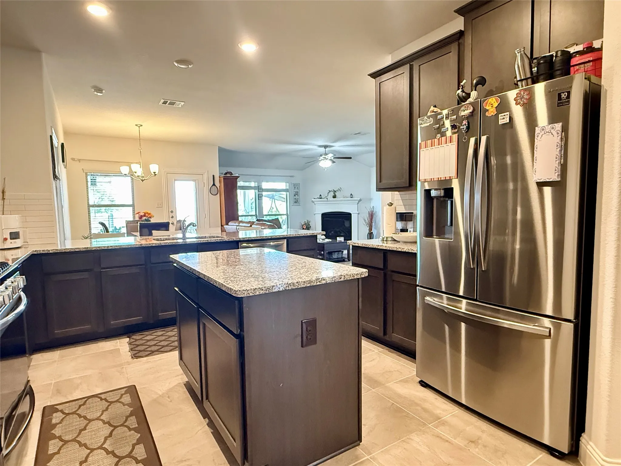 Spacious kitchen featuring an island with a light-toned granite countertop, dark wood-finish cabinetry, stainless steel appliances, and large format tile flooring