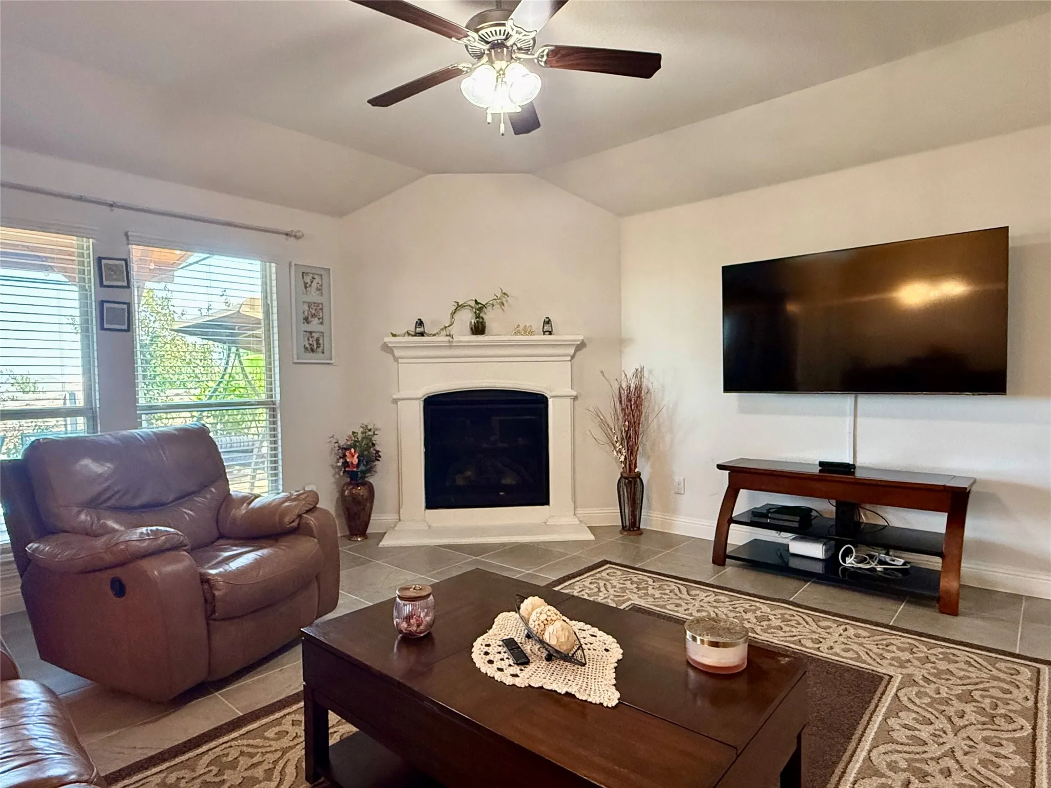 Living area featuring a corner fireplace with a white mantel, ceiling fan, large windows, and tile flooring