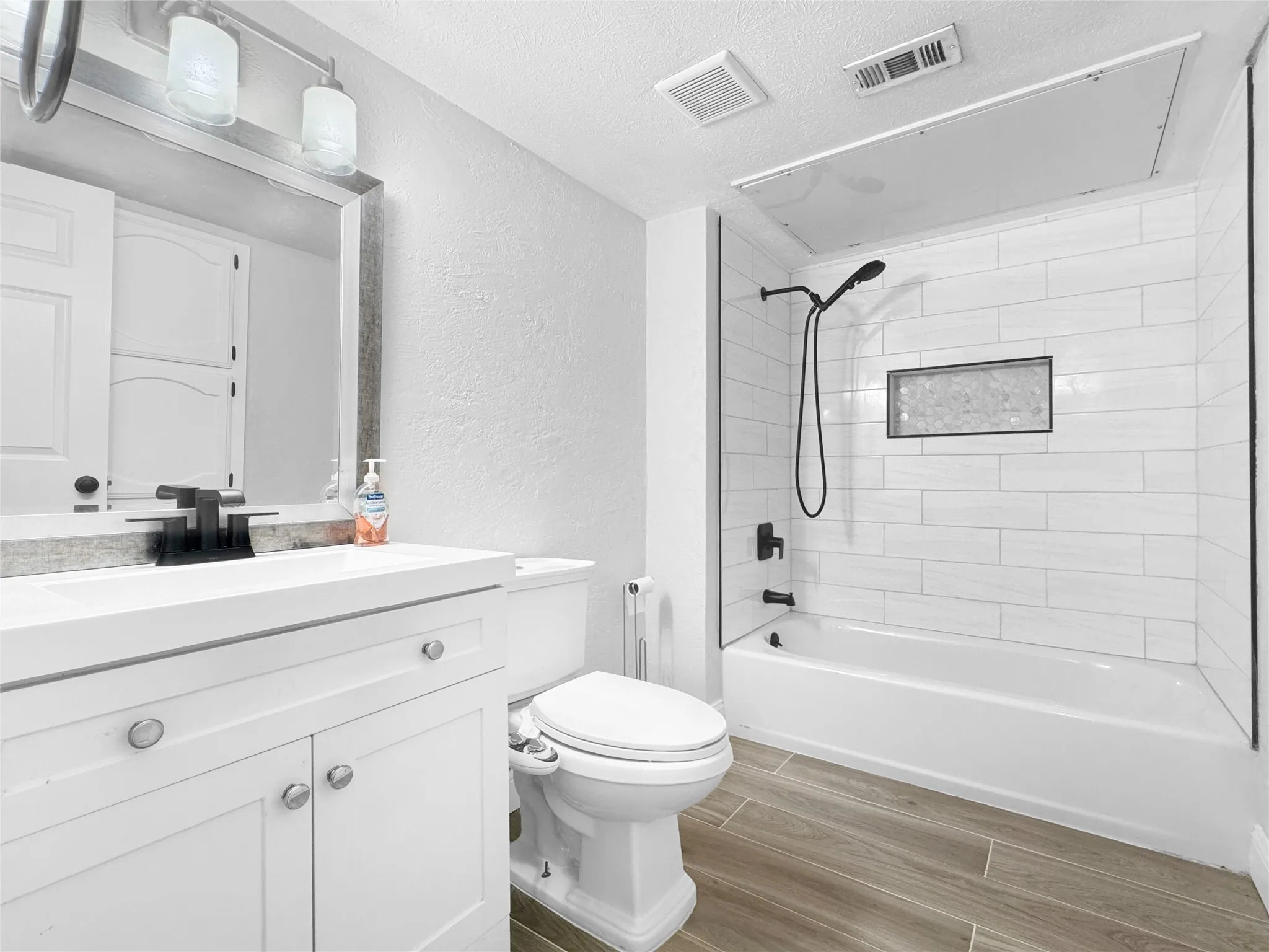 Bathroom featuring a white vanity with integrated sink, black fixtures, wood-finish flooring, and a tub/shower combination with subway tile surround and built-in niche