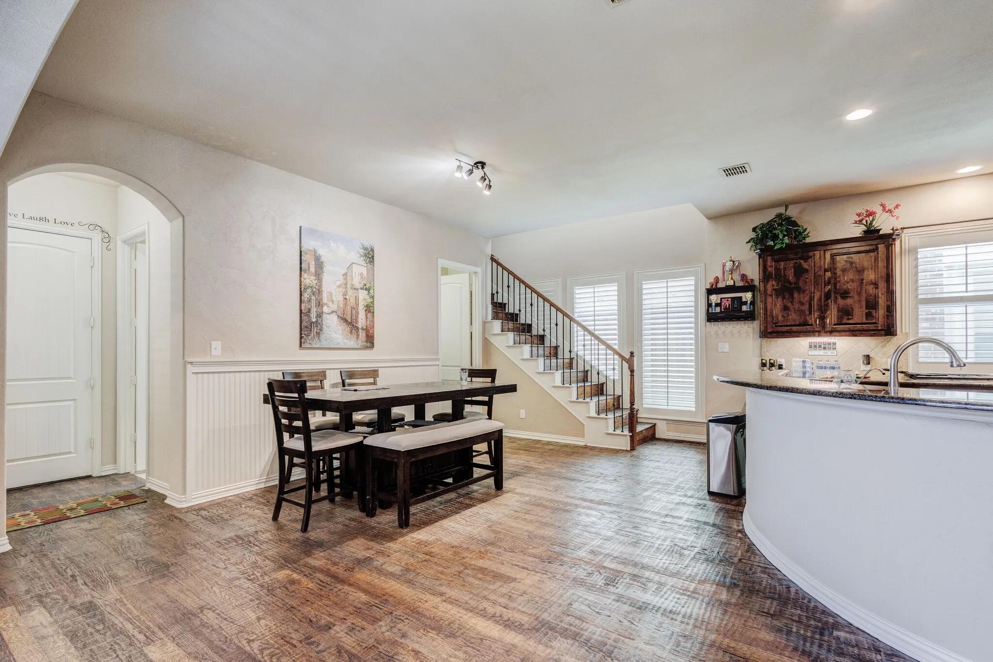 Open concept living area featuring wood-finish flooring, a staircase with dark wood handrails and white balusters, and white plantation shutters