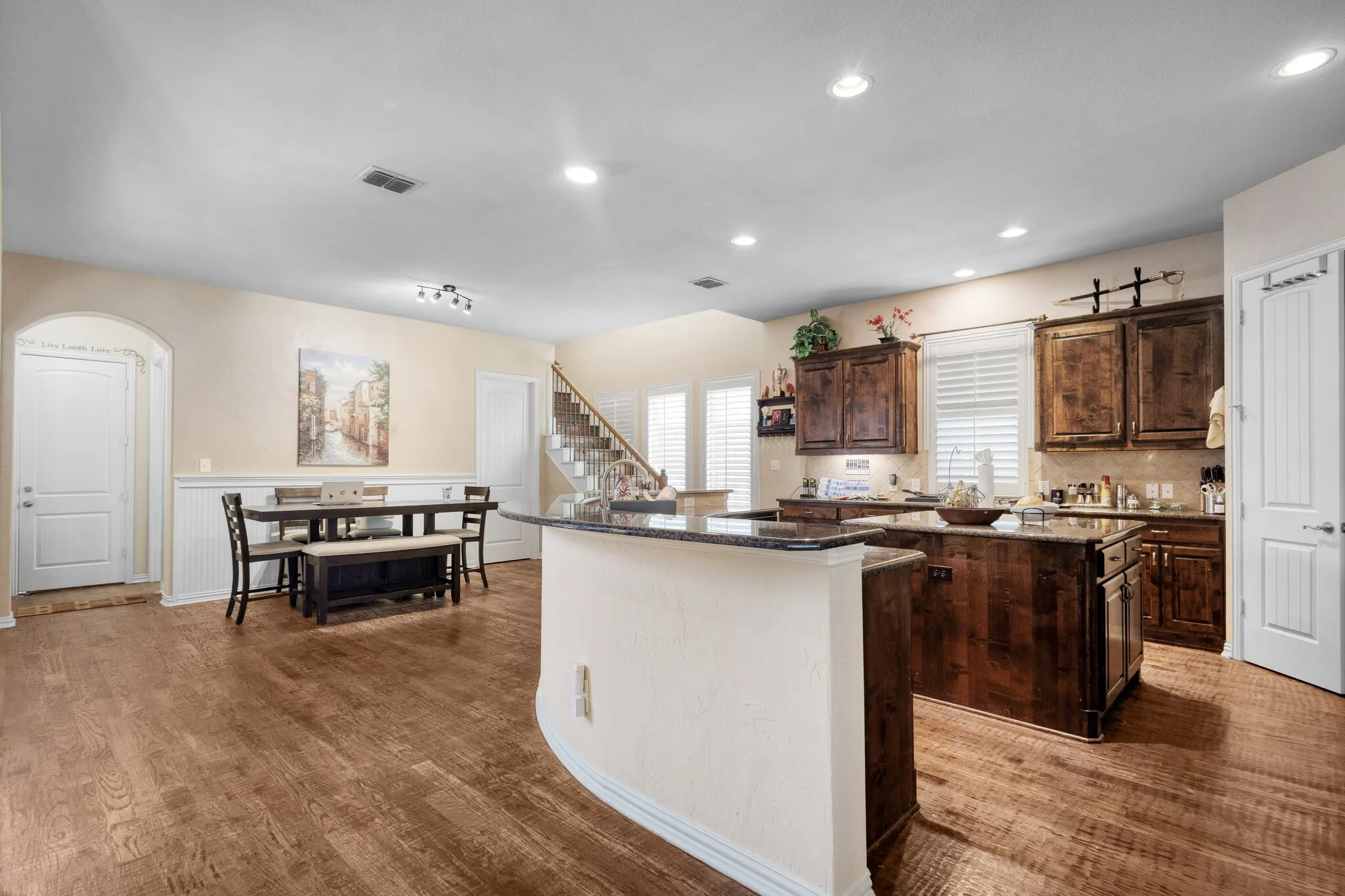 Open-concept living space featuring wood-finish flooring, a kitchen with dark wood cabinetry and granite countertops, and a staircase with white risers
