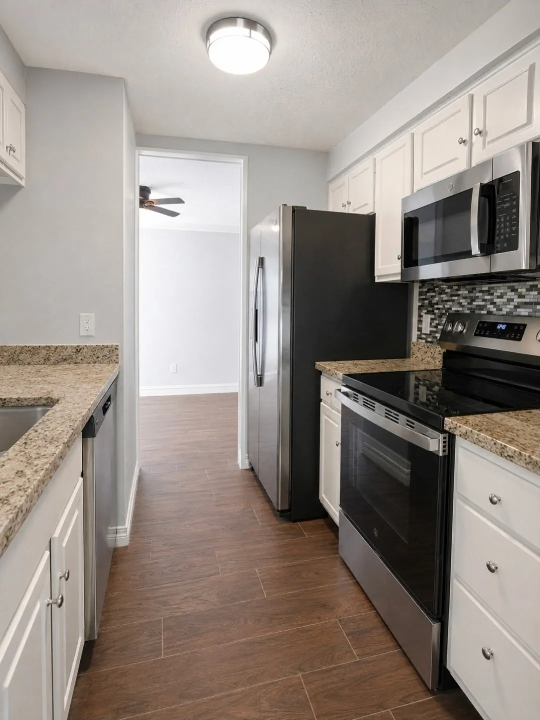 Galley kitchen featuring wood-finish flooring, white cabinetry, granite-patterned countertops, a mosaic tile backsplash, and stainless steel appliances