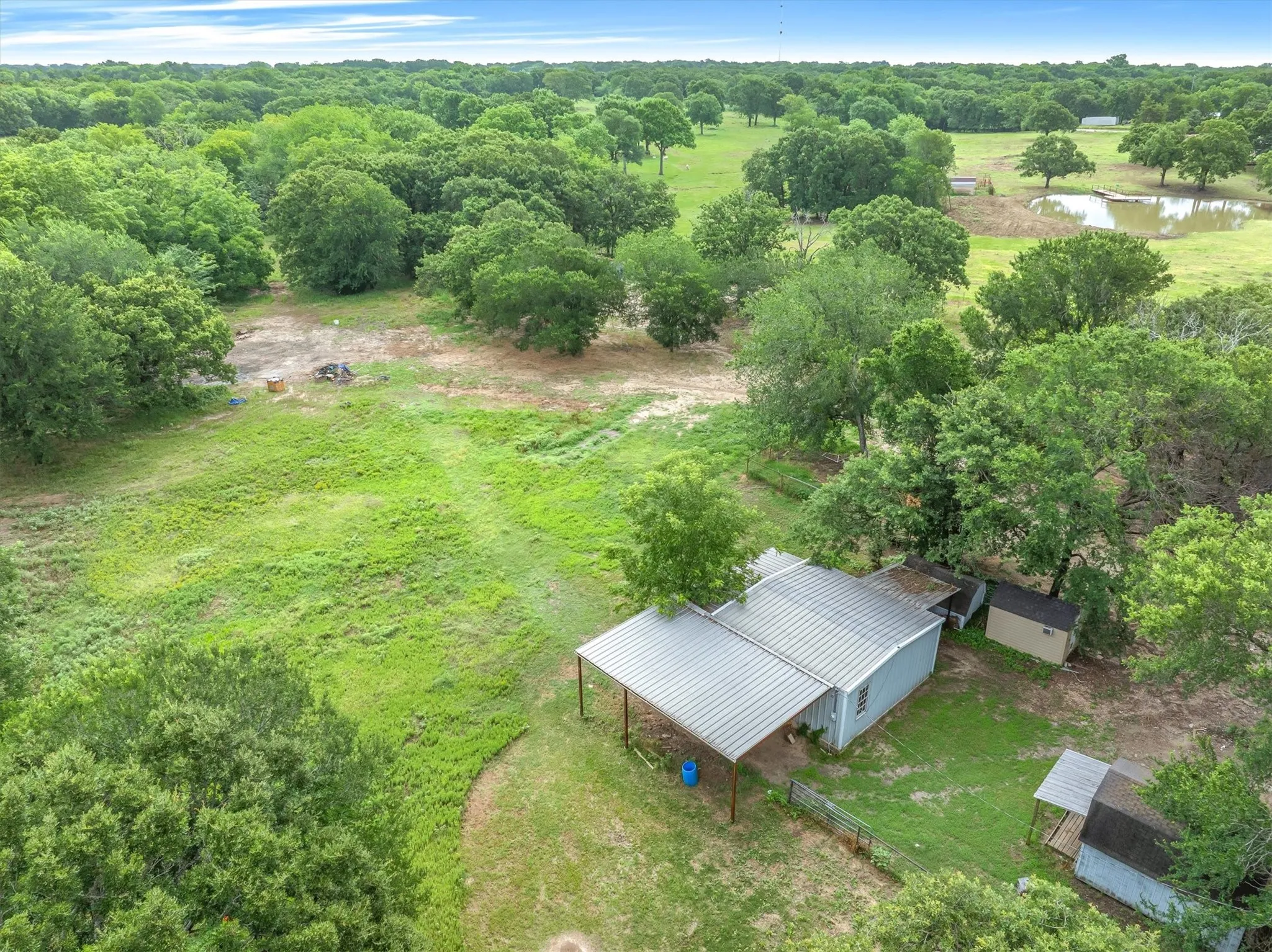 Expansive acreage featuring abundant mature trees, a visible pond, multiple outbuildings, and a main structure with a metal roof and covered porch