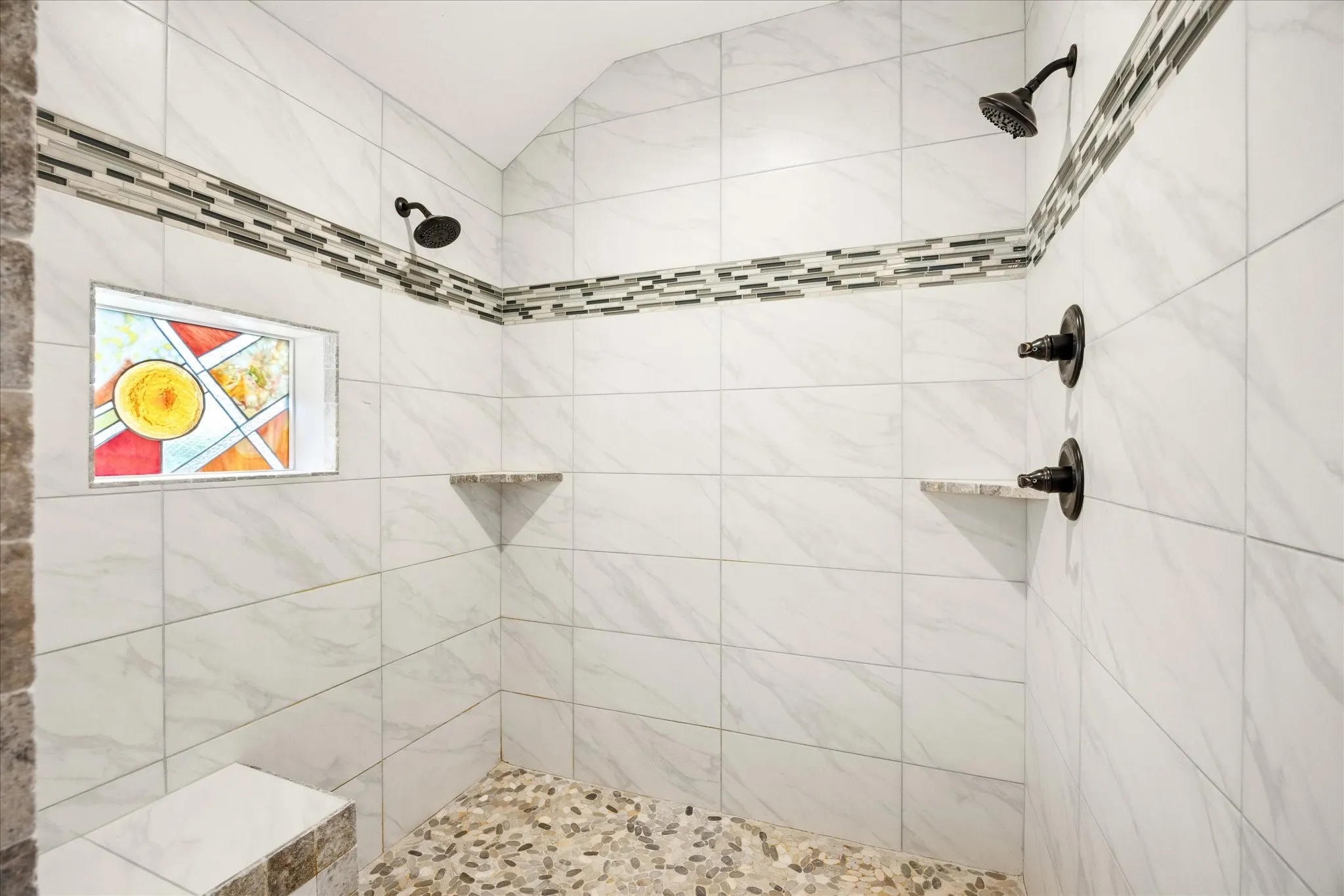 Walk-in shower featuring a pebble-stone floor, dual shower heads with oil-rubbed bronze fixtures, a built-in bench, two corner shelves, and a decorative stained glass window