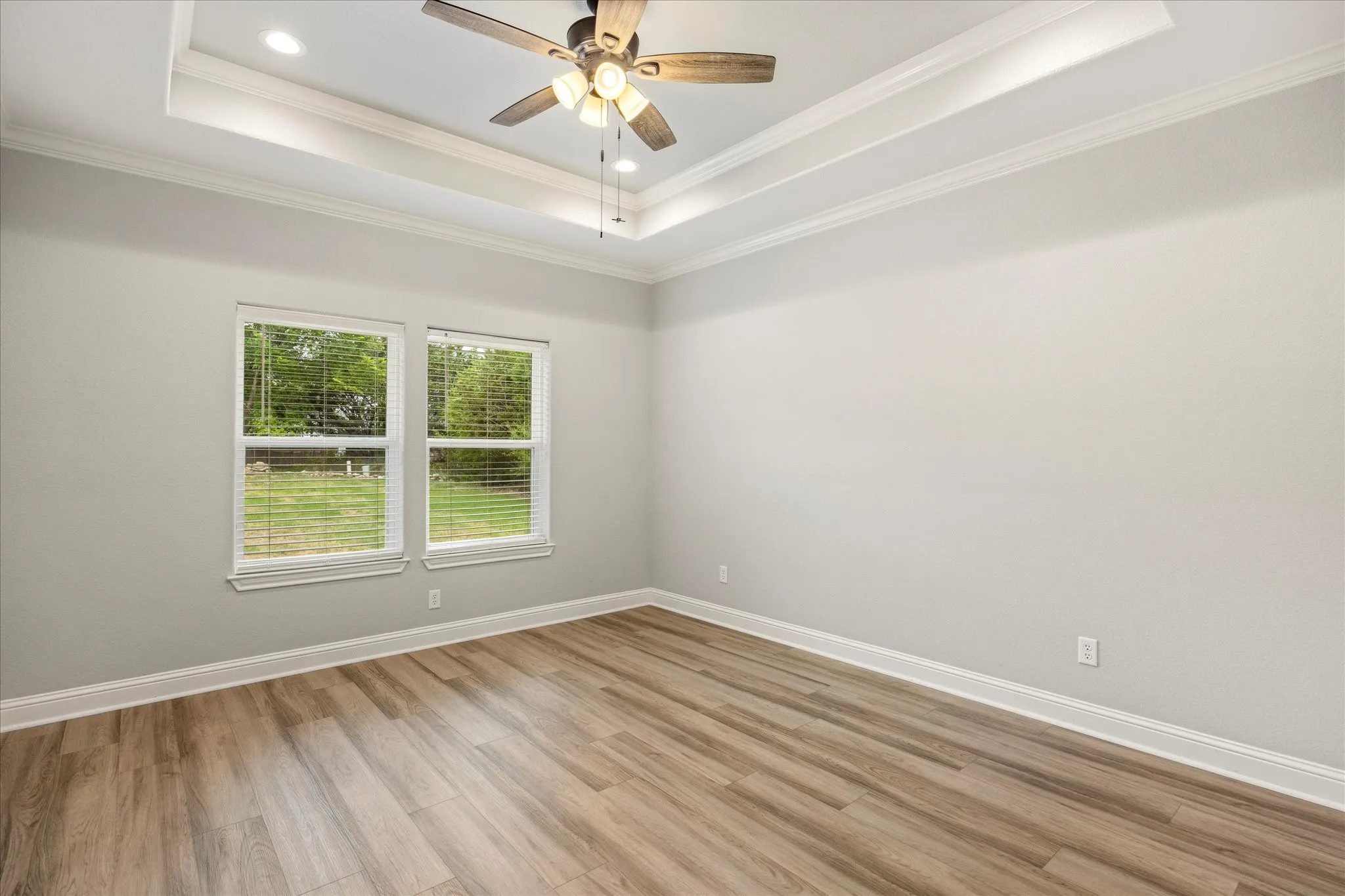 Spacious room featuring wood-finish flooring, two large windows with blinds, a tray ceiling with recessed lighting, and a ceiling fan