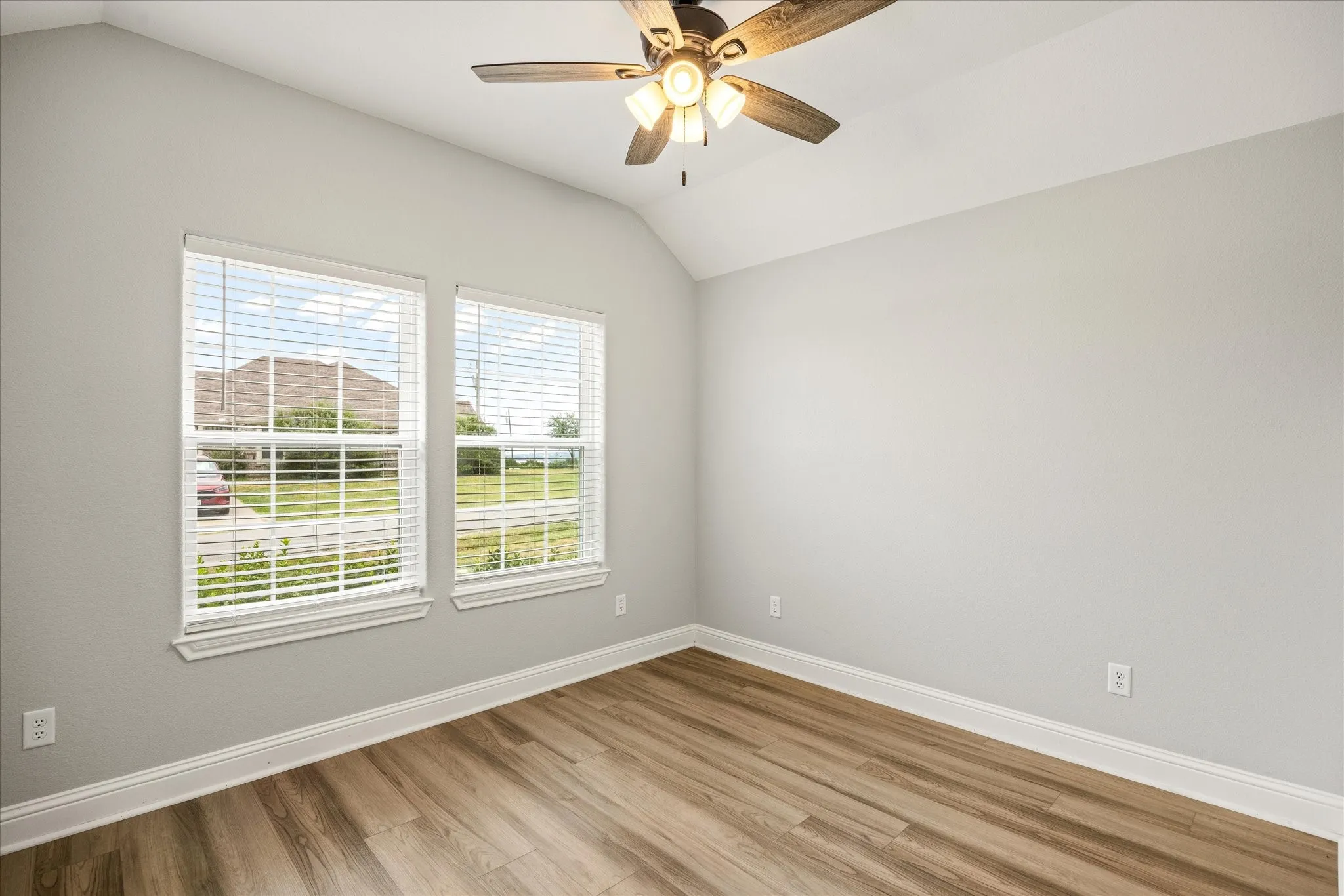 Room with a vaulted ceiling, wood-finish flooring, and two windows featuring blinds