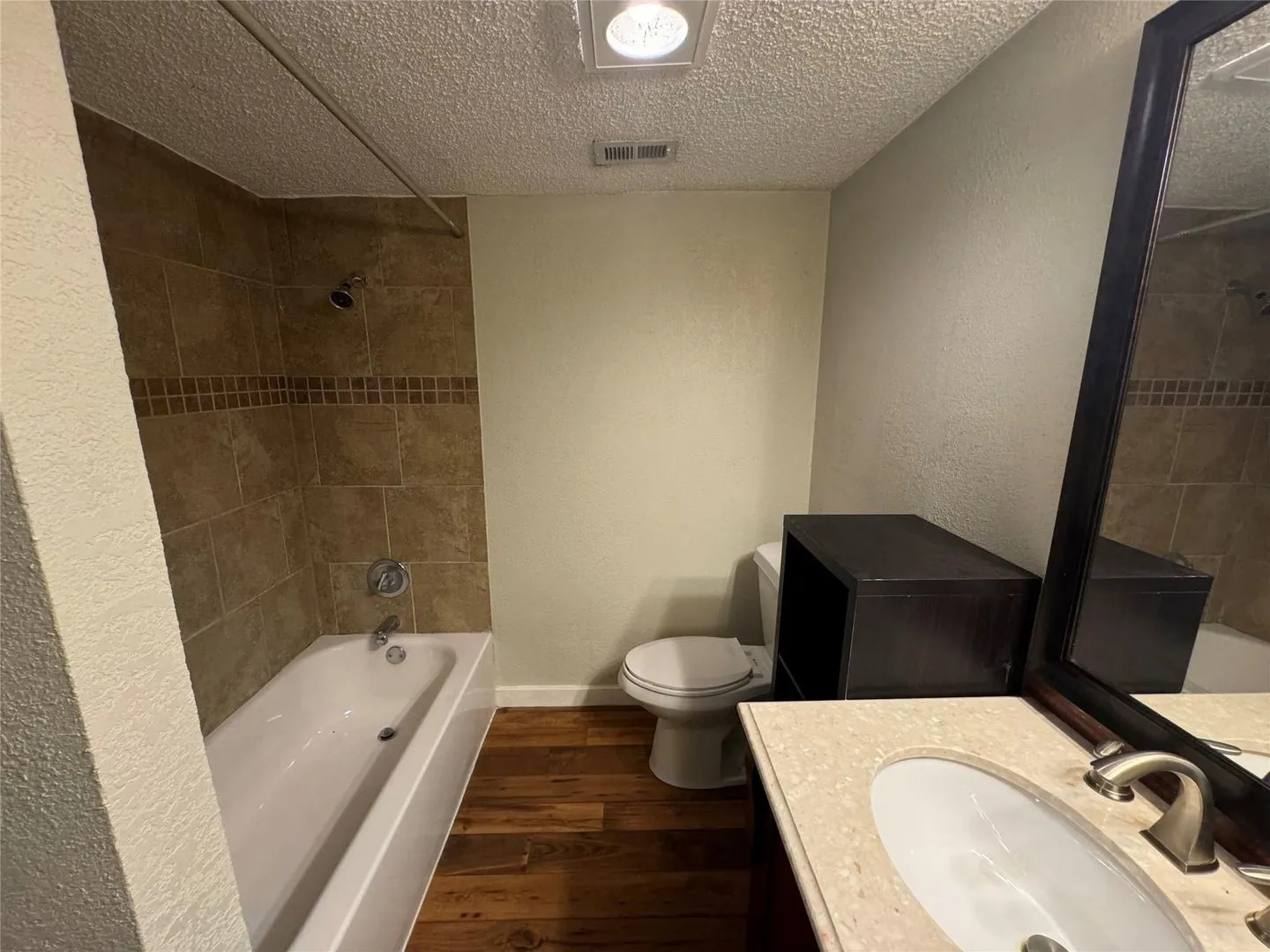 Bathroom featuring a porcelain tub with tiled surround, wood-finish flooring, a vanity with an oval sink and light-toned countertop, and a dark framed mirror