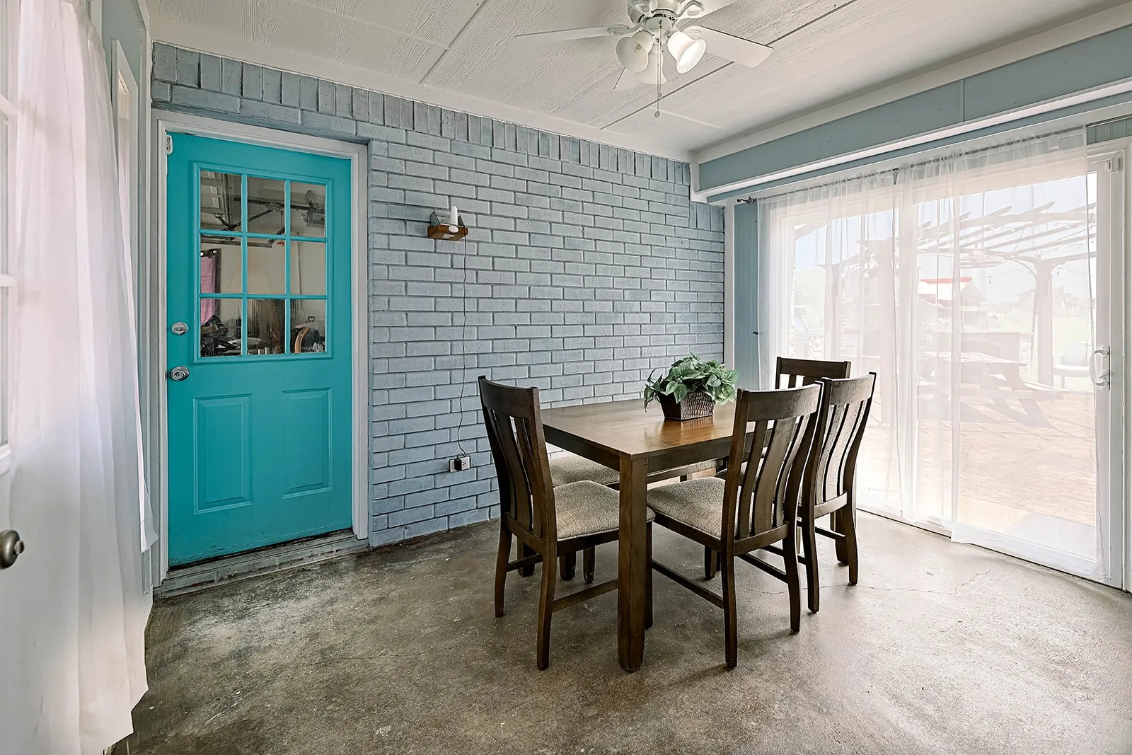 Enclosed patio featuring a turquoise paneled door with windowpanes, light blue brick-finish walls, a ceiling fan with light fixture, and concrete flooring