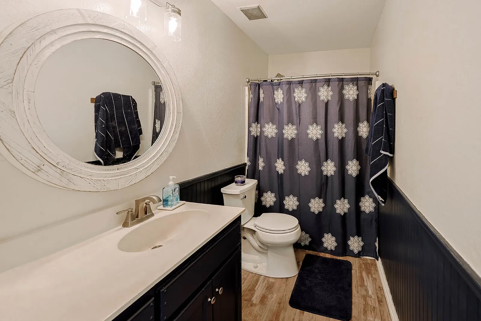 Bathroom featuring wood-finish flooring, a white vanity countertop with an integrated sink, a dark wood-finish vanity cabinet, a round white-framed mirror, and a toilet
