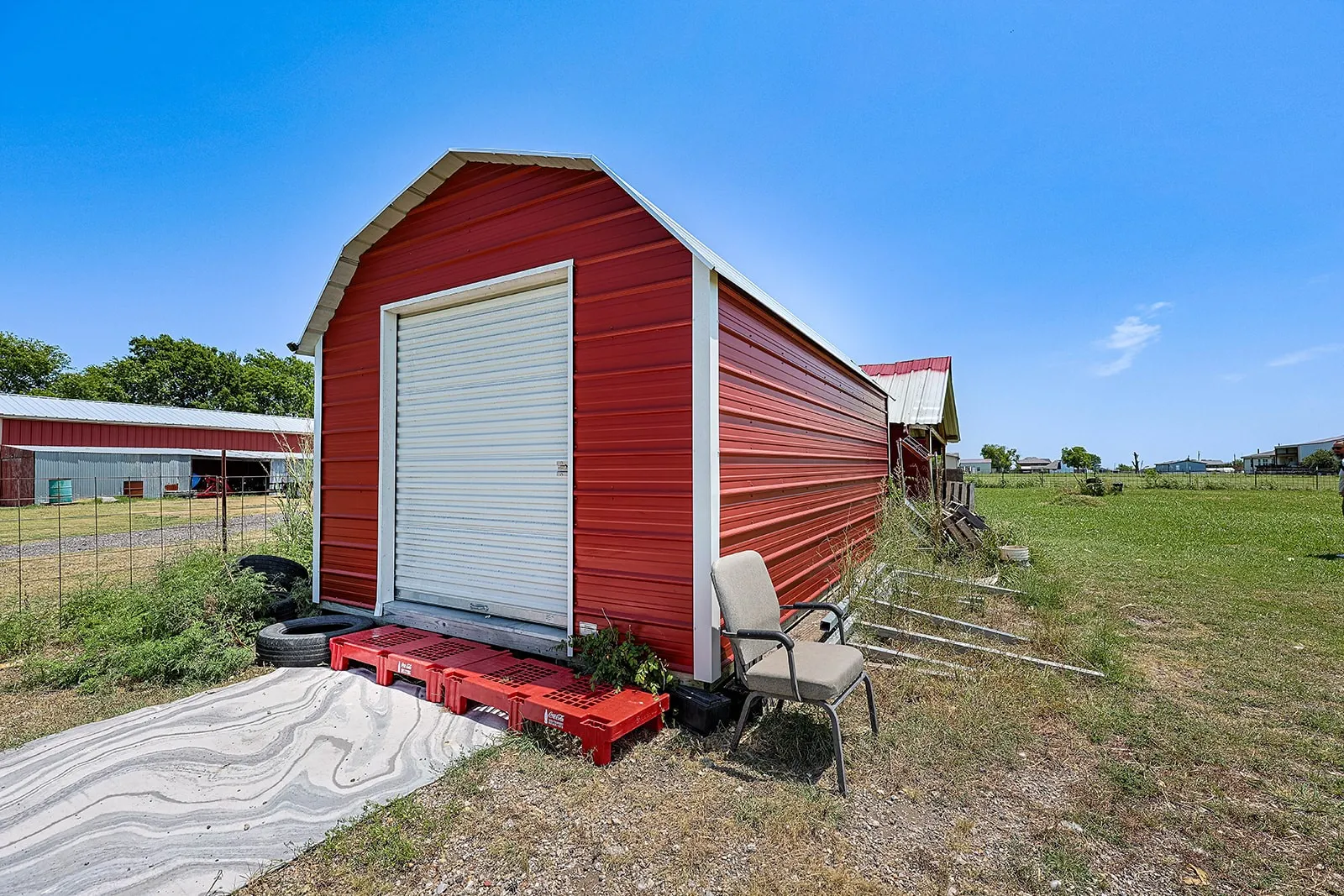 Red metal storage building featuring a white roll-up door, a concrete entry ramp, a gable roofline, and visible corrugated siding