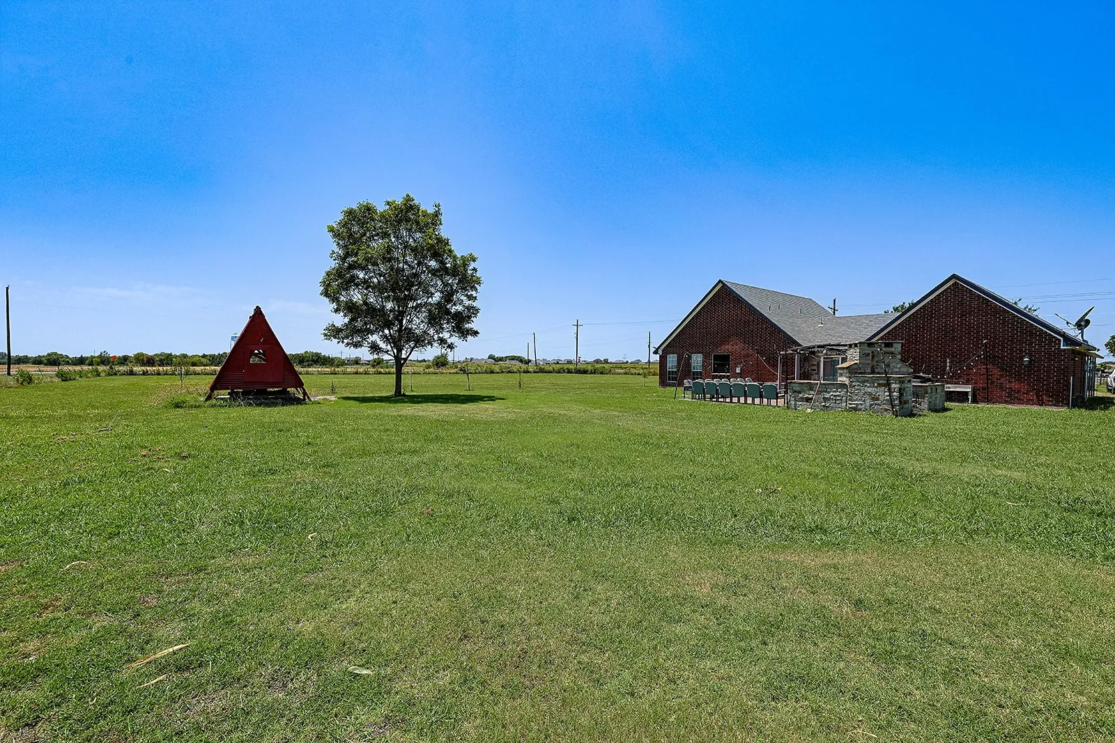 Expansive grassy yard with mature tree and a brick exterior dwelling featuring a patio area with a stone fireplace
