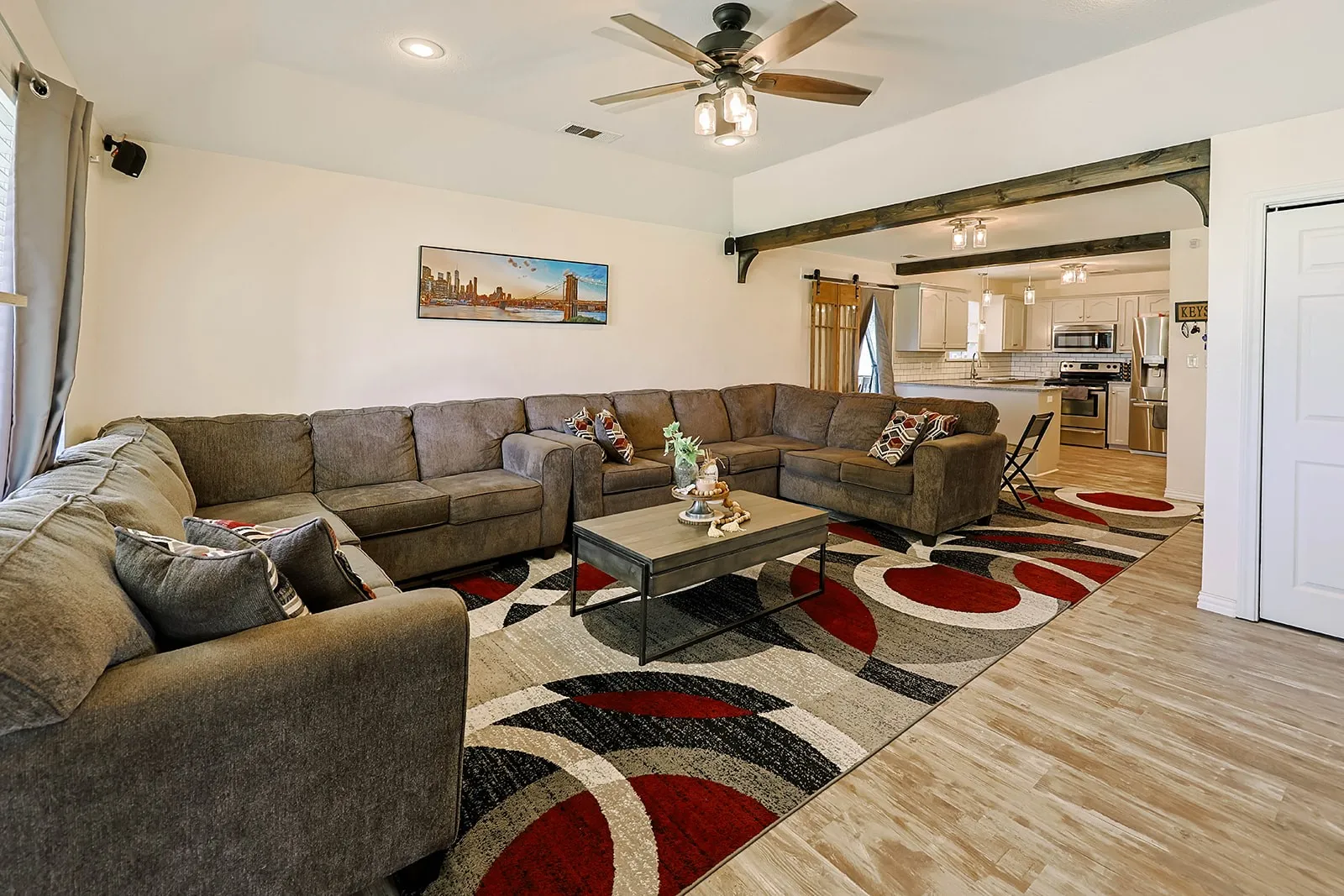 Open-concept living space featuring wood-finish flooring, a ceiling fan with integrated lighting, recessed lighting, and decorative wood beams defining the entry to the kitchen