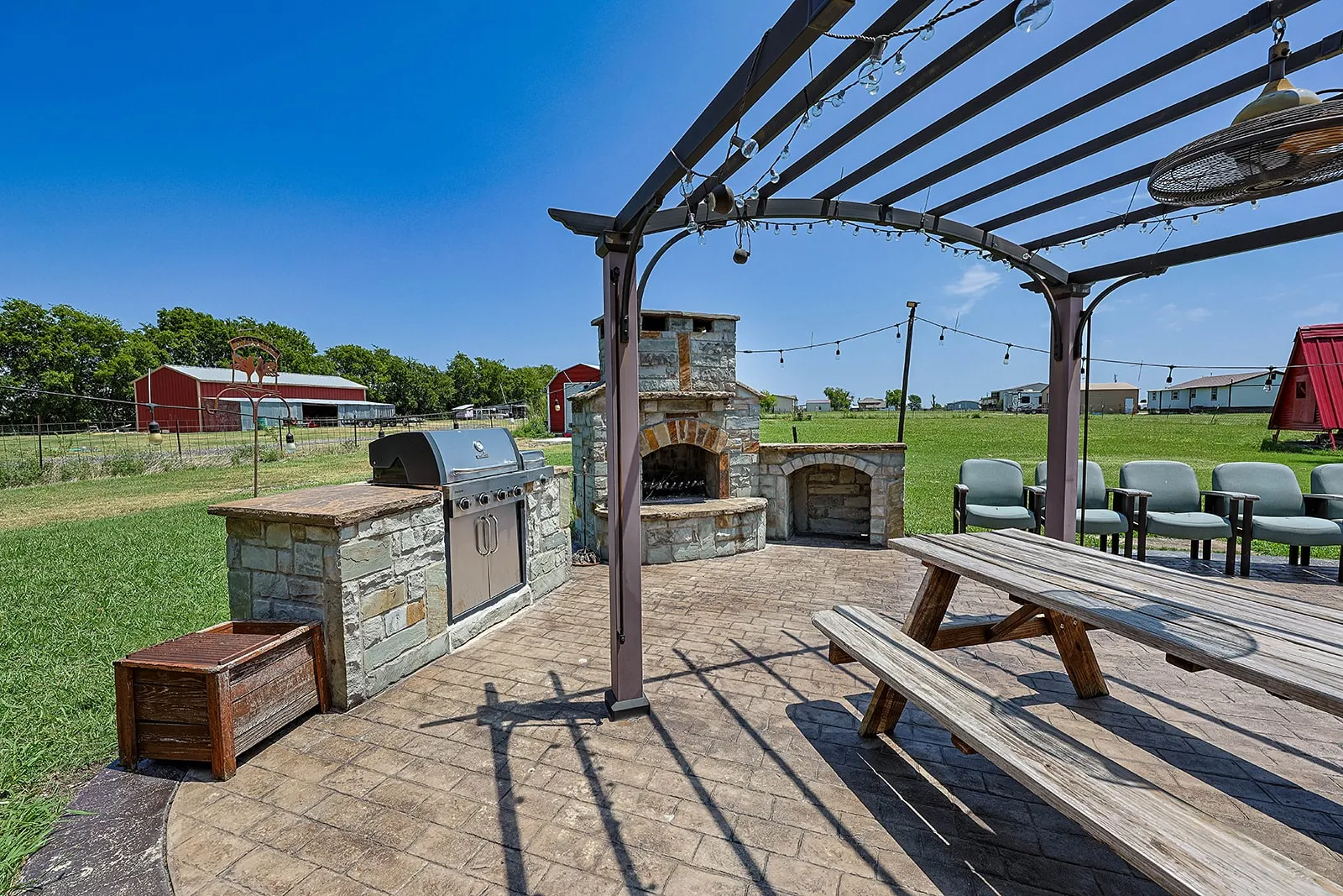Stamped concrete patio featuring an outdoor kitchen with built-in grill and stone base, a stone fireplace, and a metal pergola with string lighting