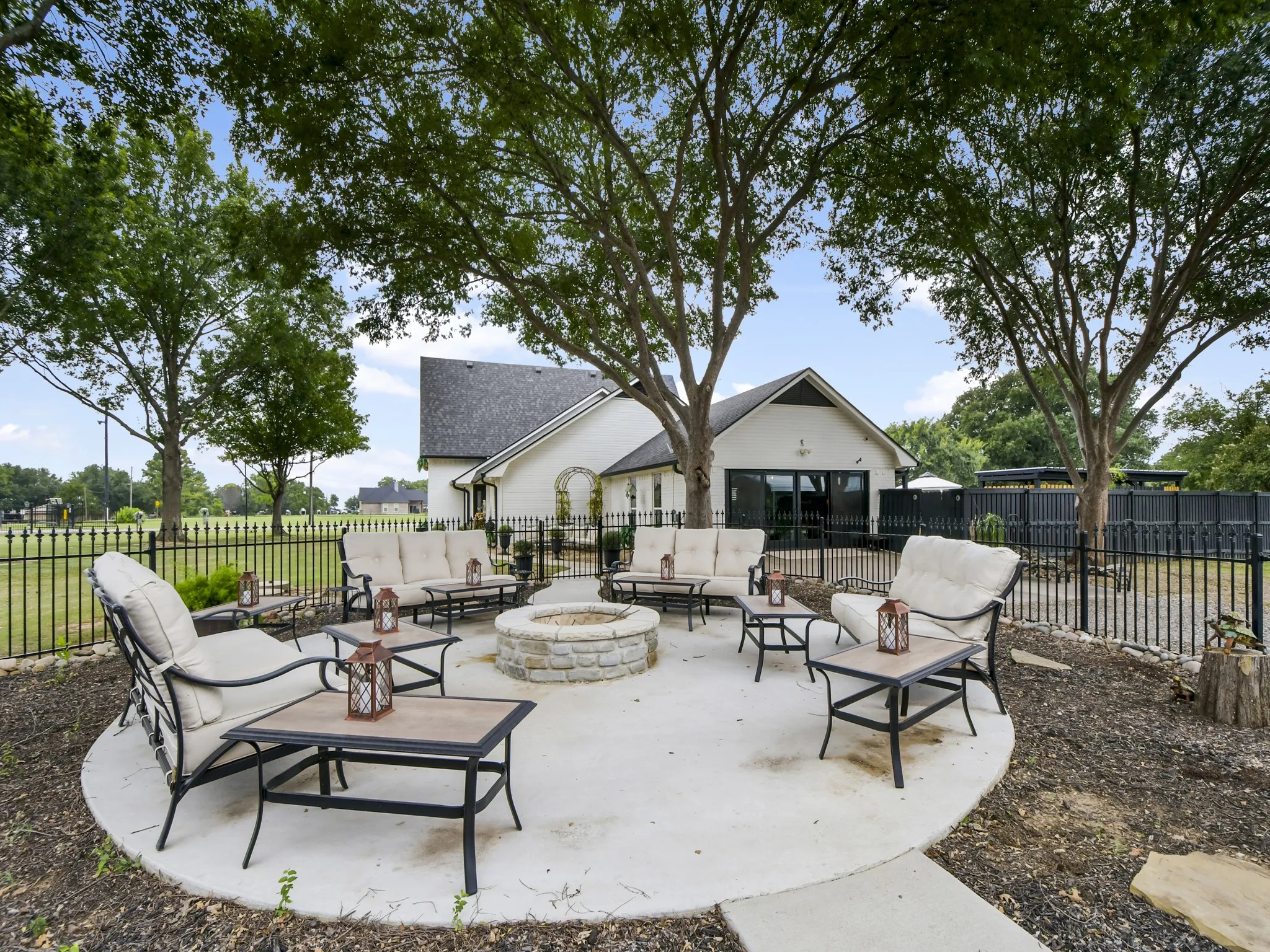 Circular concrete patio featuring a stone-clad fire pit, mature trees, and a black wrought iron fence
