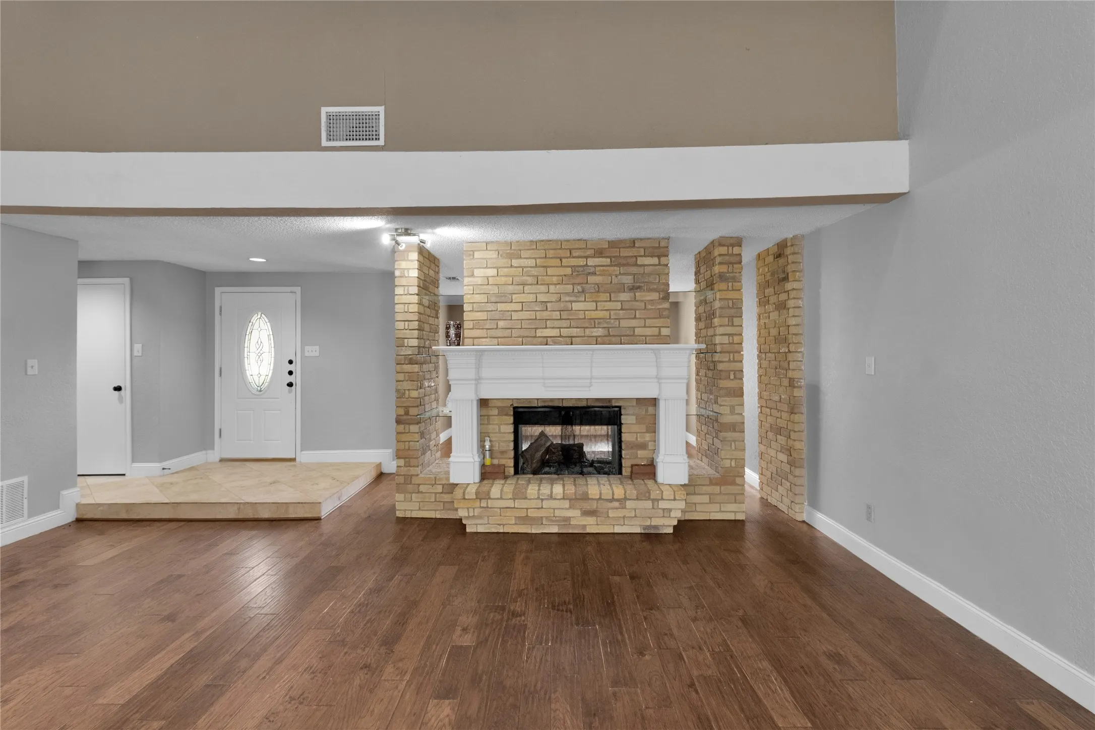 Wood-finish flooring, brick fireplace with white mantel, white entry door with oval glass insert, and tiled entryway