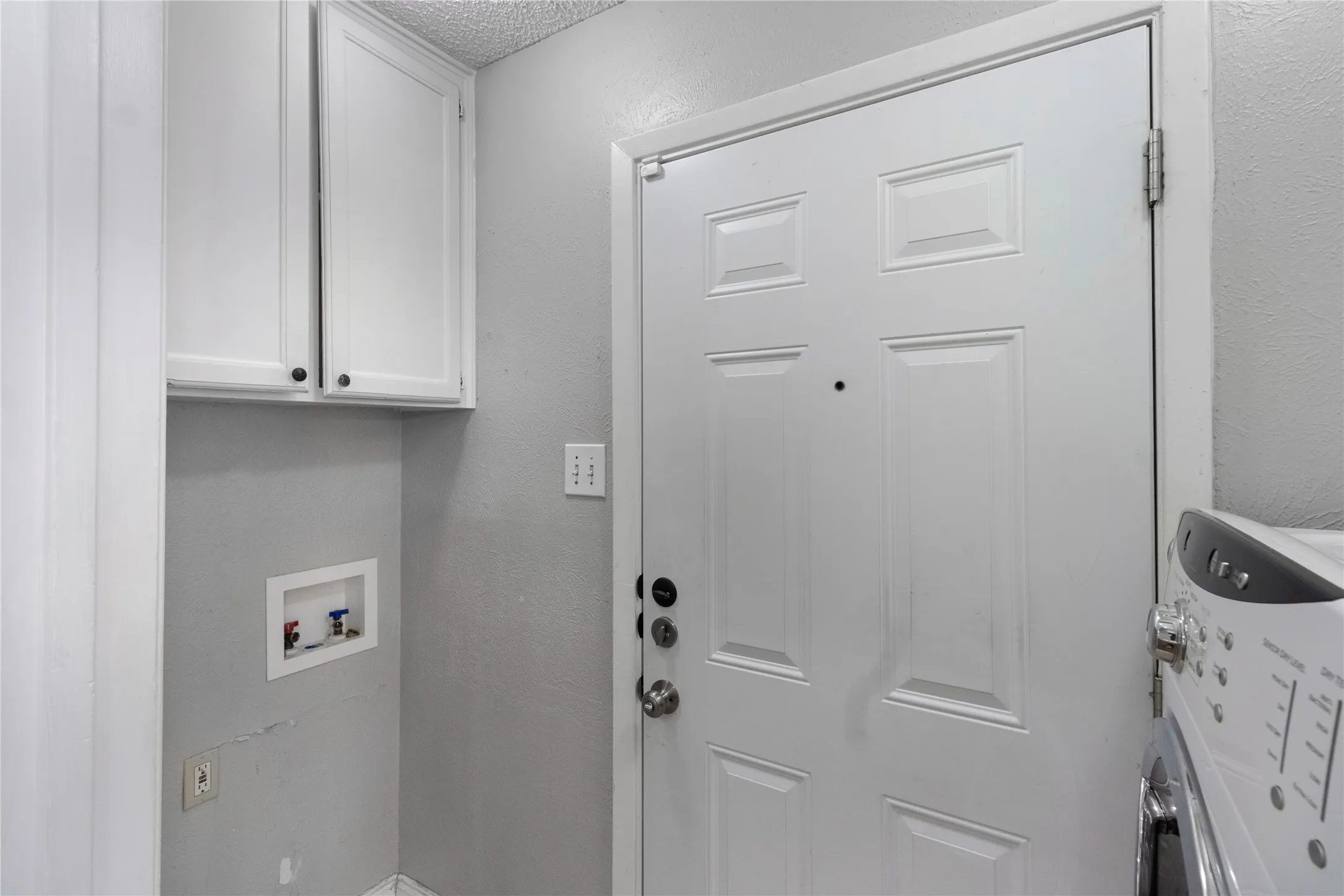 White paneled door with deadbolt, upper white cabinetry with dark hardware, and washer/dryer connections
