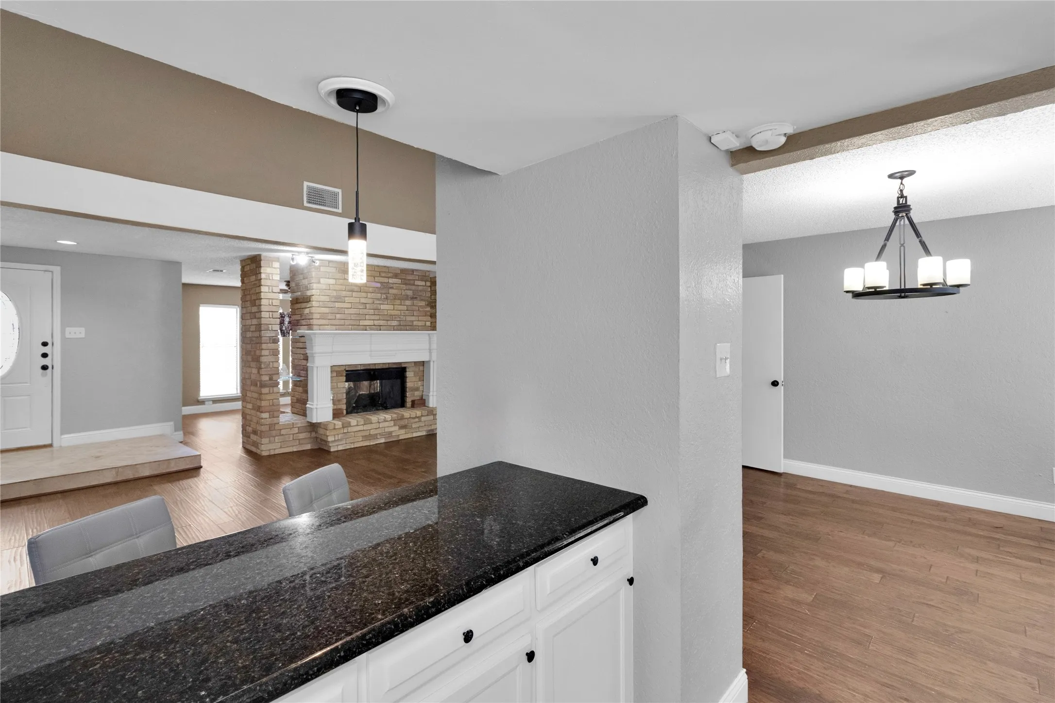 Granite-finish countertop with white cabinetry, wood-finish flooring, brick fireplace with white mantel, and a modern chandelier
