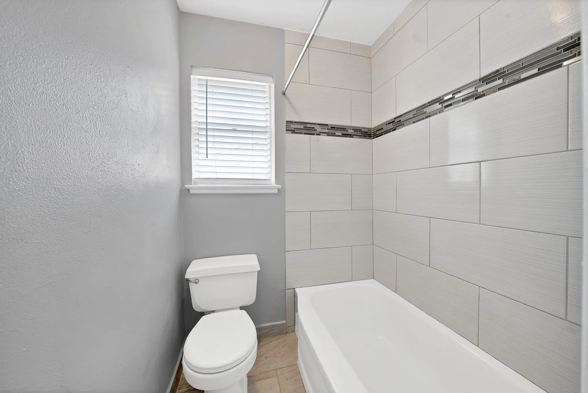 Bathroom featuring a white bathtub, light-colored subway tile surround with a mosaic accent band, and a window with blinds