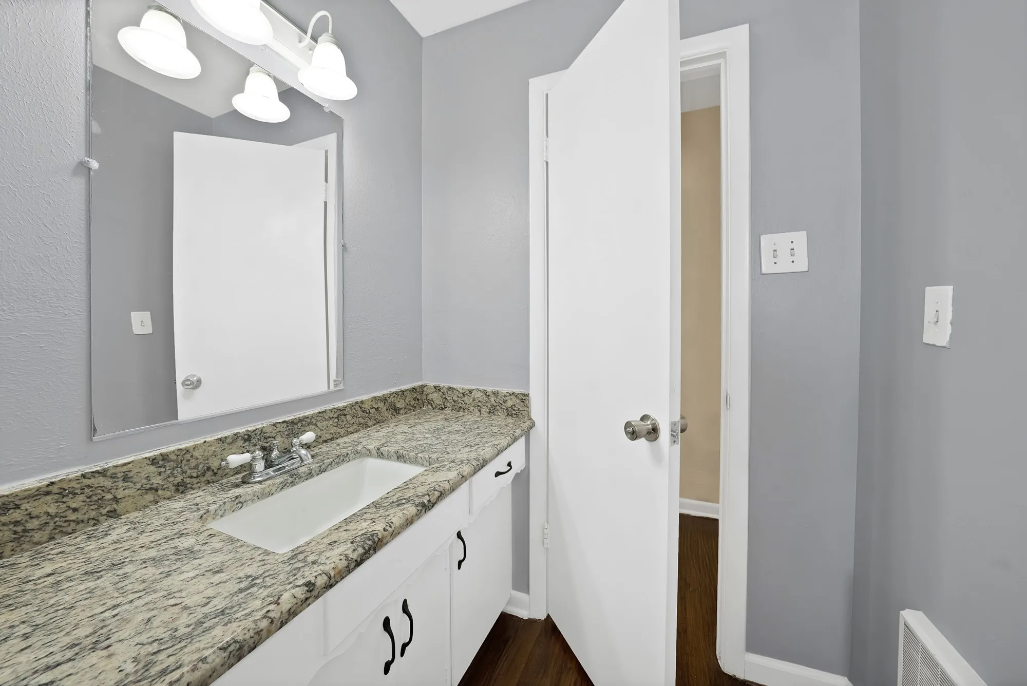 Bathroom vanity featuring an integrated sink, stone countertop, white cabinetry with black hardware, and a wall-mounted mirror with overhead lighting