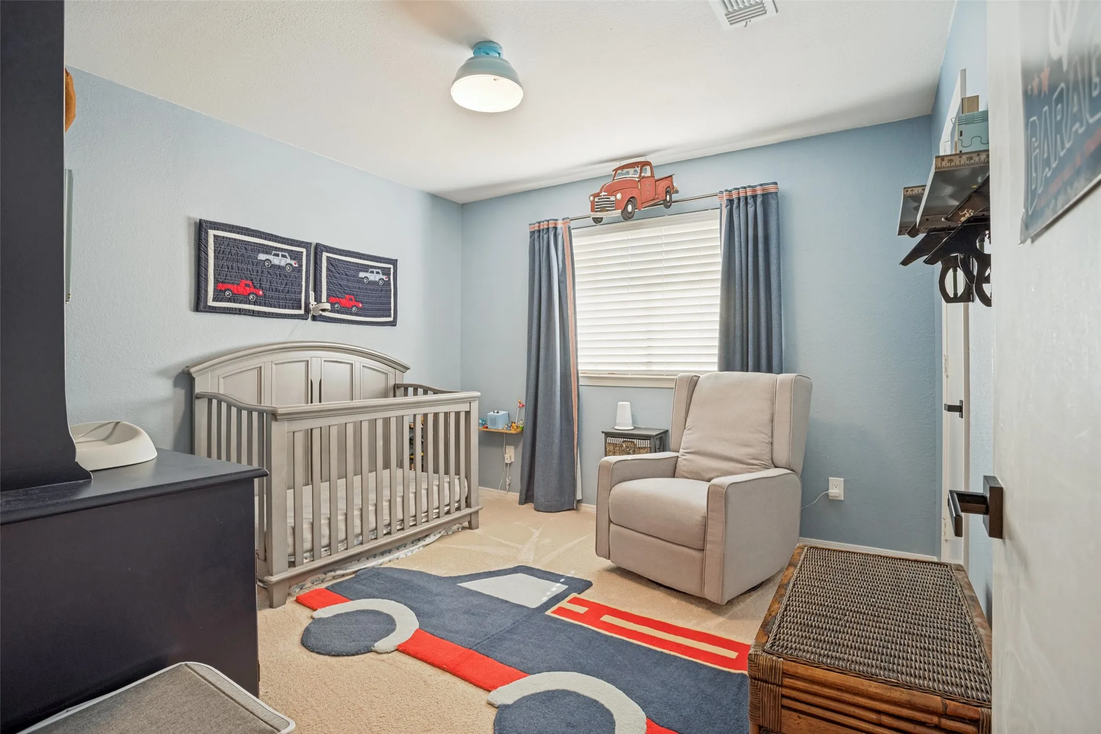 Bright nursery bedroom featuring soft blue walls, natural light from a large window with gray curtains, wood-finish flooring with coordinating area rug, wooden crib, upholstered glider chair, and ceiling-mounted fixture