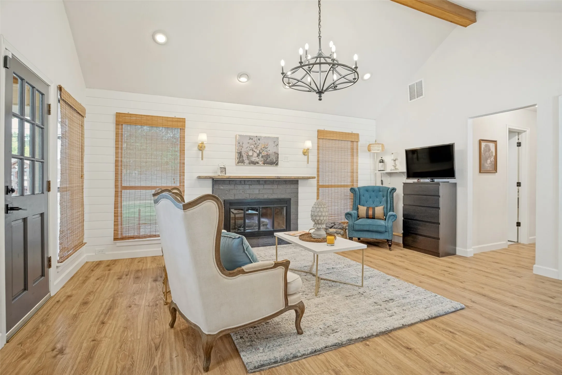 Vaulted ceiling with an exposed wood beam, wood-finish flooring, white shiplap accent wall, fireplace with stone surround, and a dark-paneled door with glass inserts