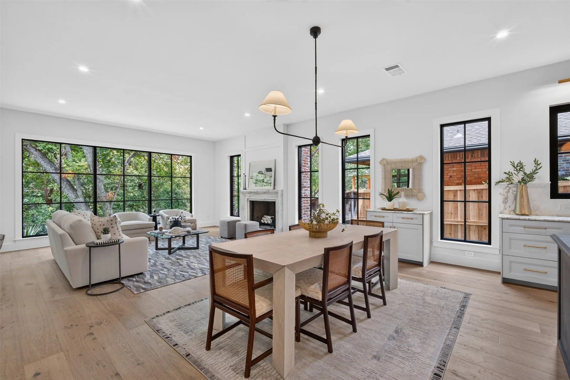 Open-concept living area featuring light wood-finish flooring, a contemporary chandelier, a white stone fireplace surround, and extensive black-framed windows