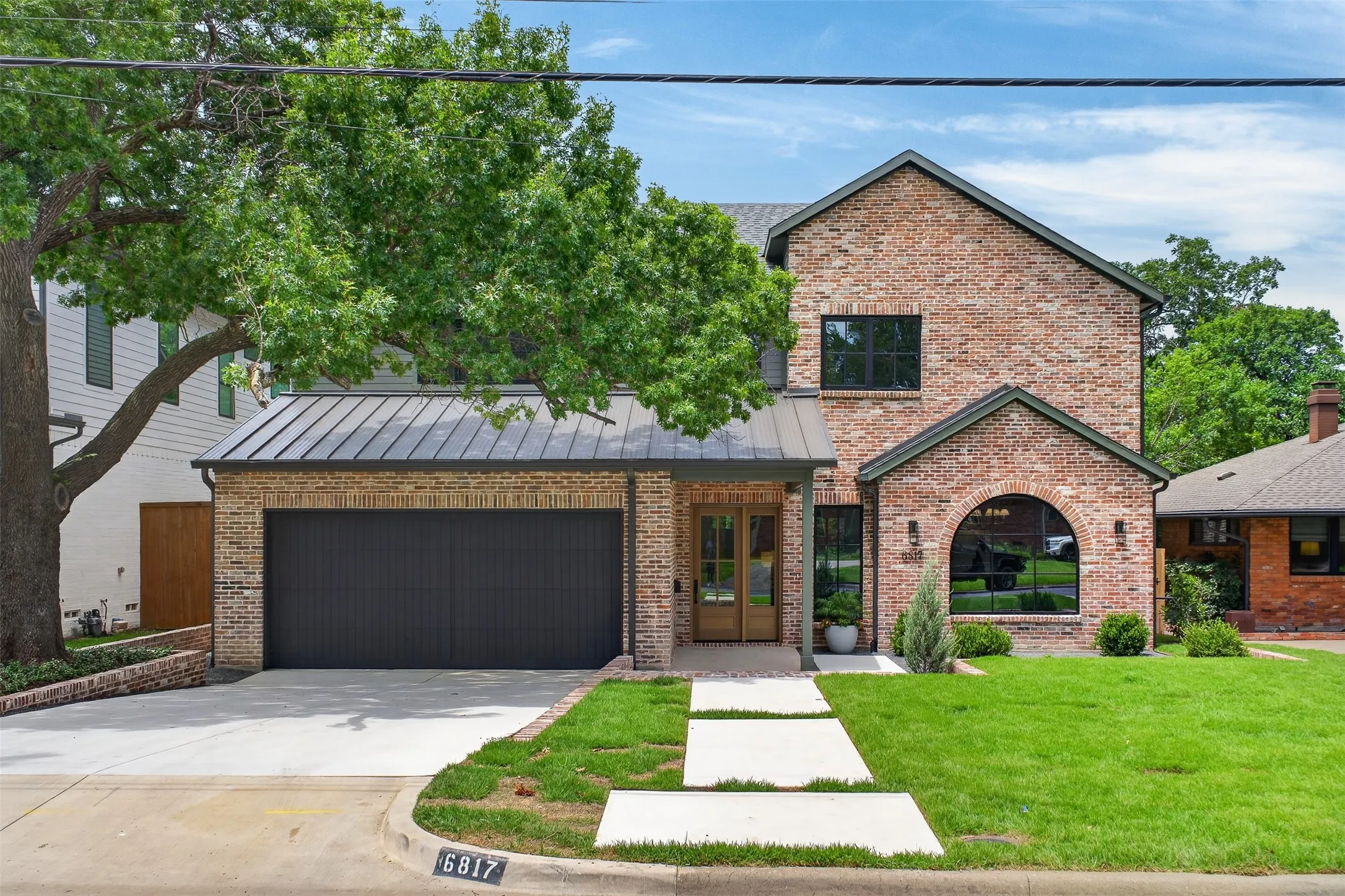 Contemporary brick exterior with a dark metal roof, a two-car garage with a dark door, a double wood-panel entrance door, and a prominent arched window feature