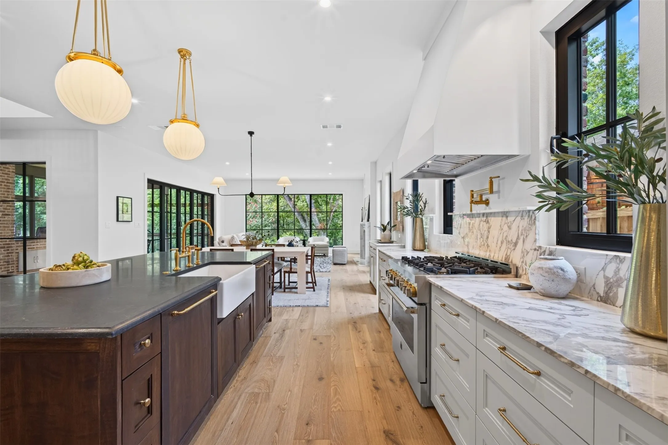 Gourmet kitchen featuring a substantial island with dark wood cabinetry and a farmhouse sink, light wood-finish flooring, light stone countertops with a full-height backsplash, a professional-grade range, and abundant natural light from numerous windows