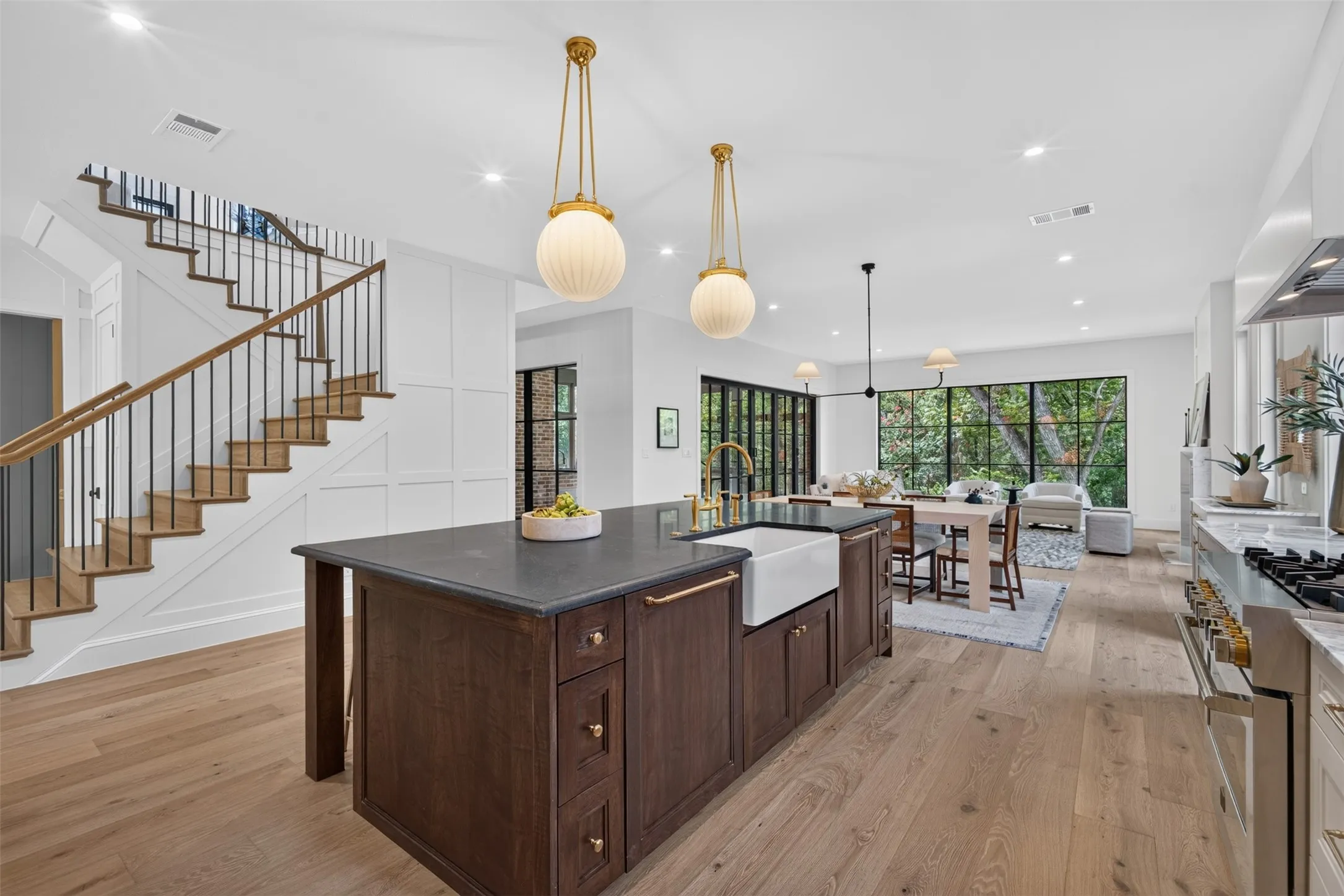 Open-concept living space featuring a dark wood kitchen island with an integrated farmhouse sink, light wood-finish flooring, and a contemporary staircase with dark iron balusters