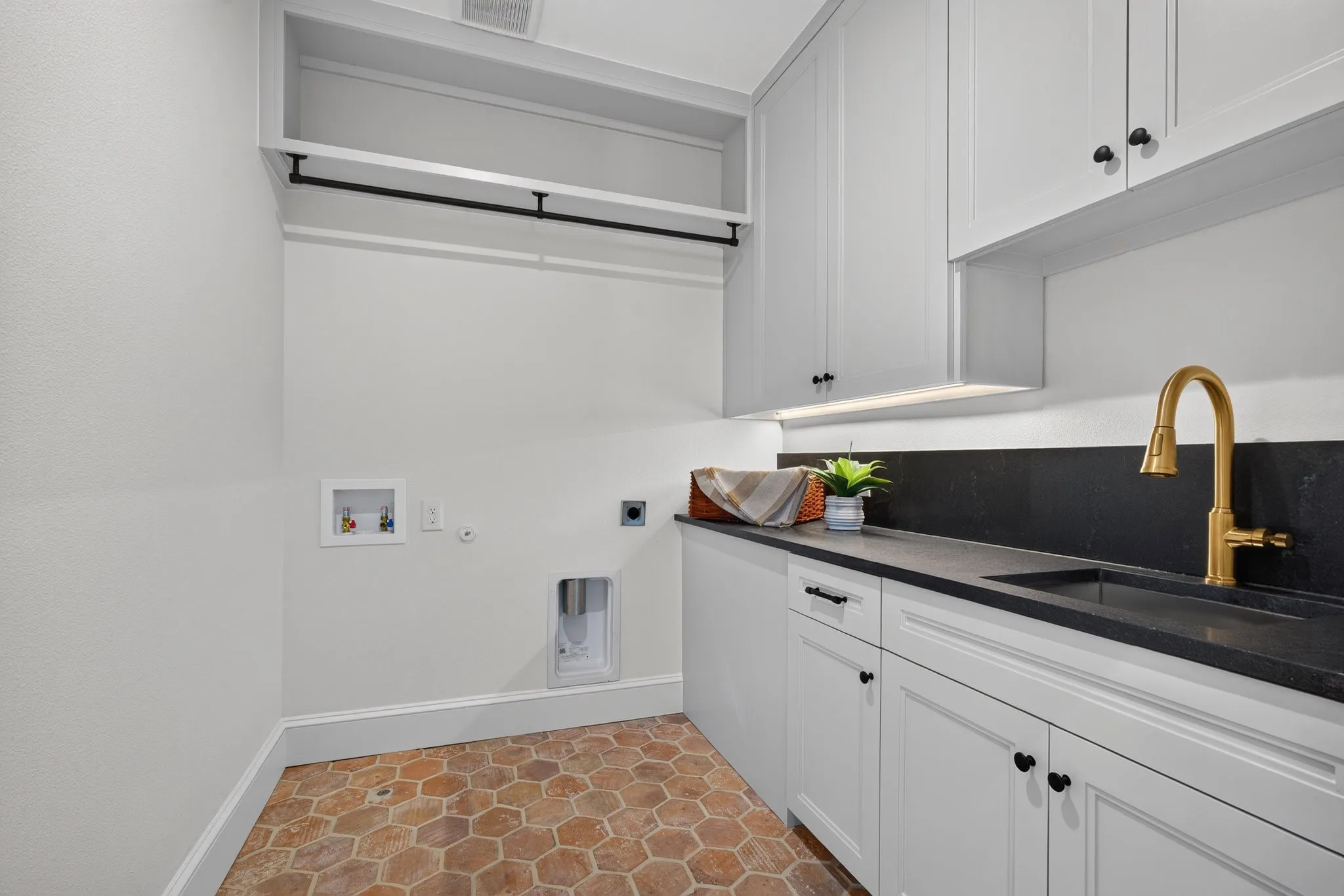 Laundry room featuring hexagonal tile flooring, white shaker cabinetry with black hardware, black countertop with integrated sink, and a brass-finish faucet