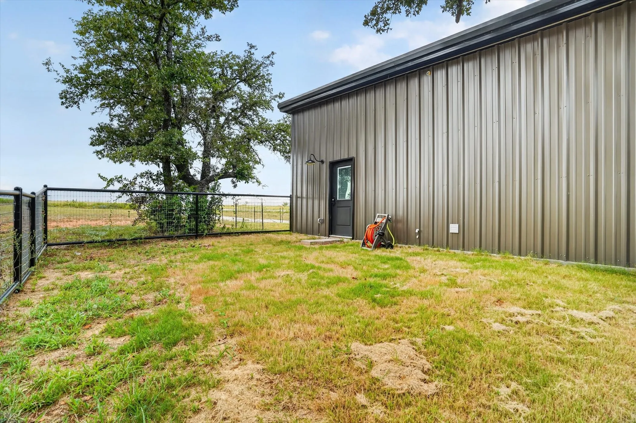 Corrugated metal siding structure with a black entry door, a metal roof, and a perimeter fence enclosing a natural grass area
