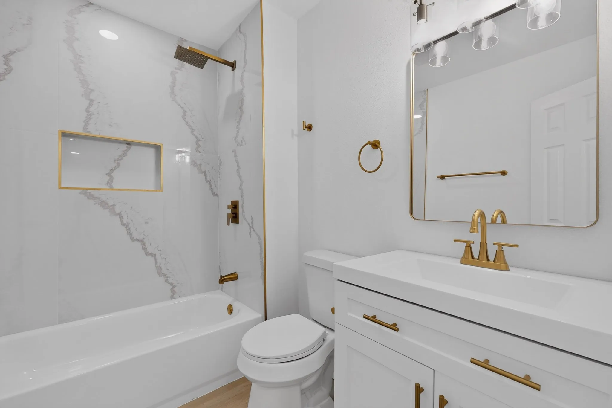Bathroom featuring a white vanity with gold-tone hardware, a rectangular mirror with integrated lighting, a white bathtub, a tiled shower surround with a gold-tone shower head, and a built-in shower niche
