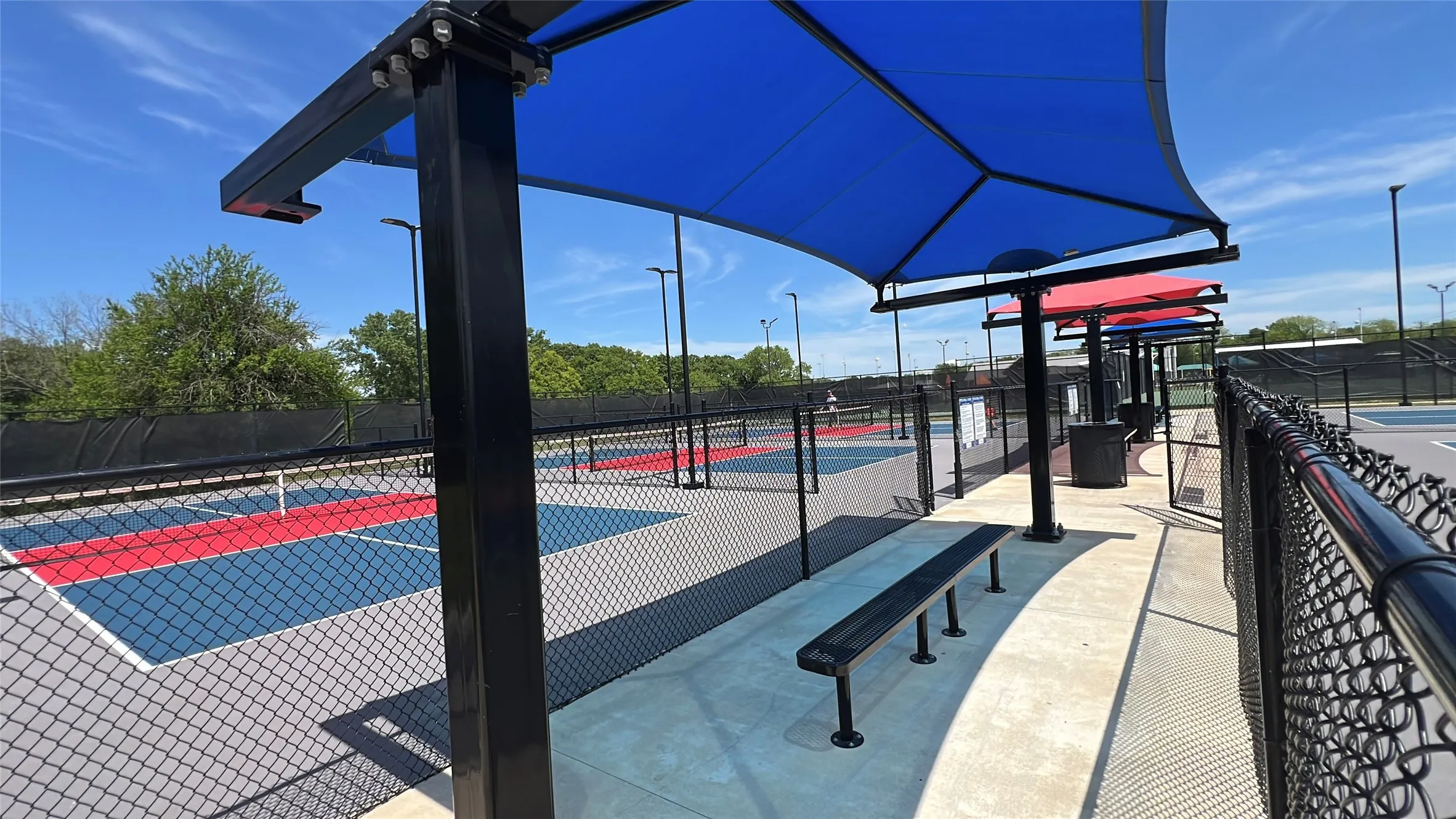 Outdoor sports court area featuring multiple courts with blue and red playing surfaces, black chain-link fencing, and overhead lighting