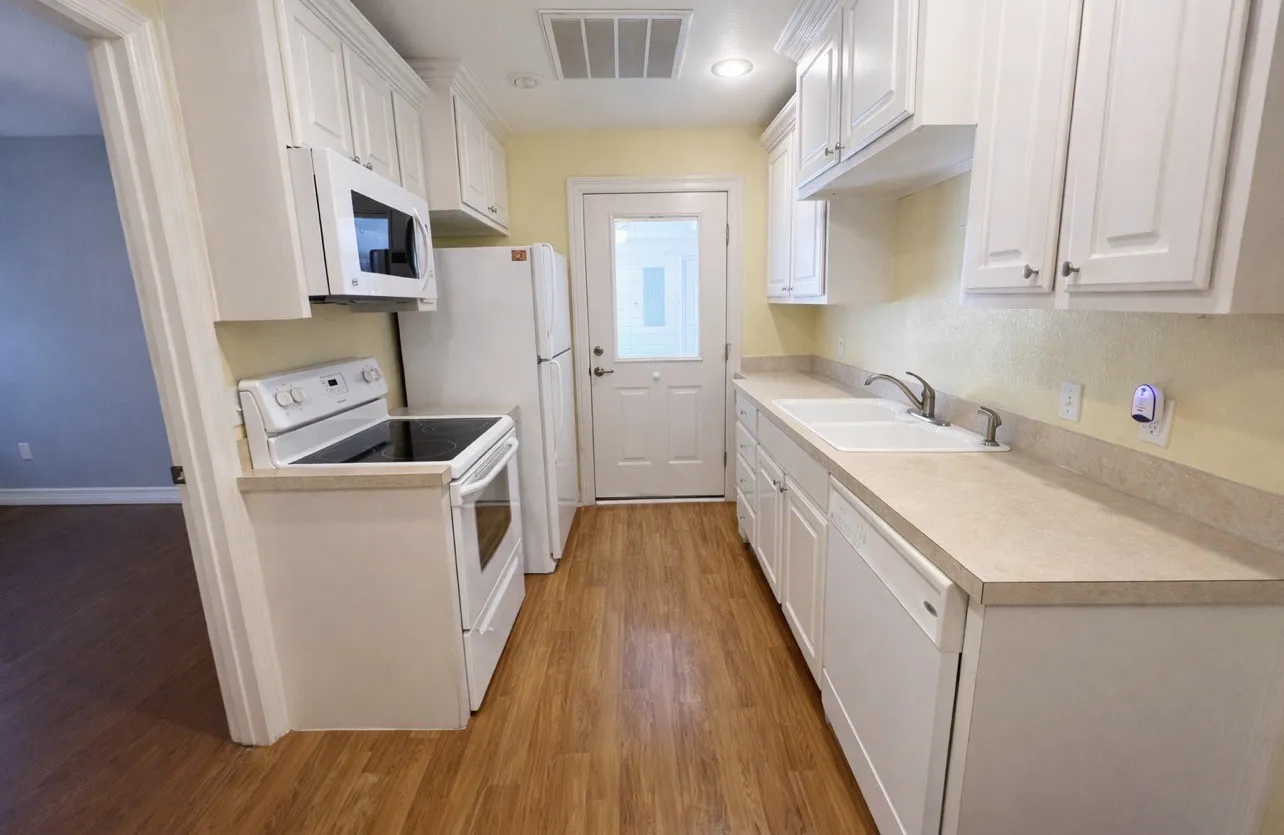 Galley kitchen featuring wood-finish flooring, white cabinetry, light-toned countertops, and a full suite of white appliances including an electric range, microwave, and refrigerator